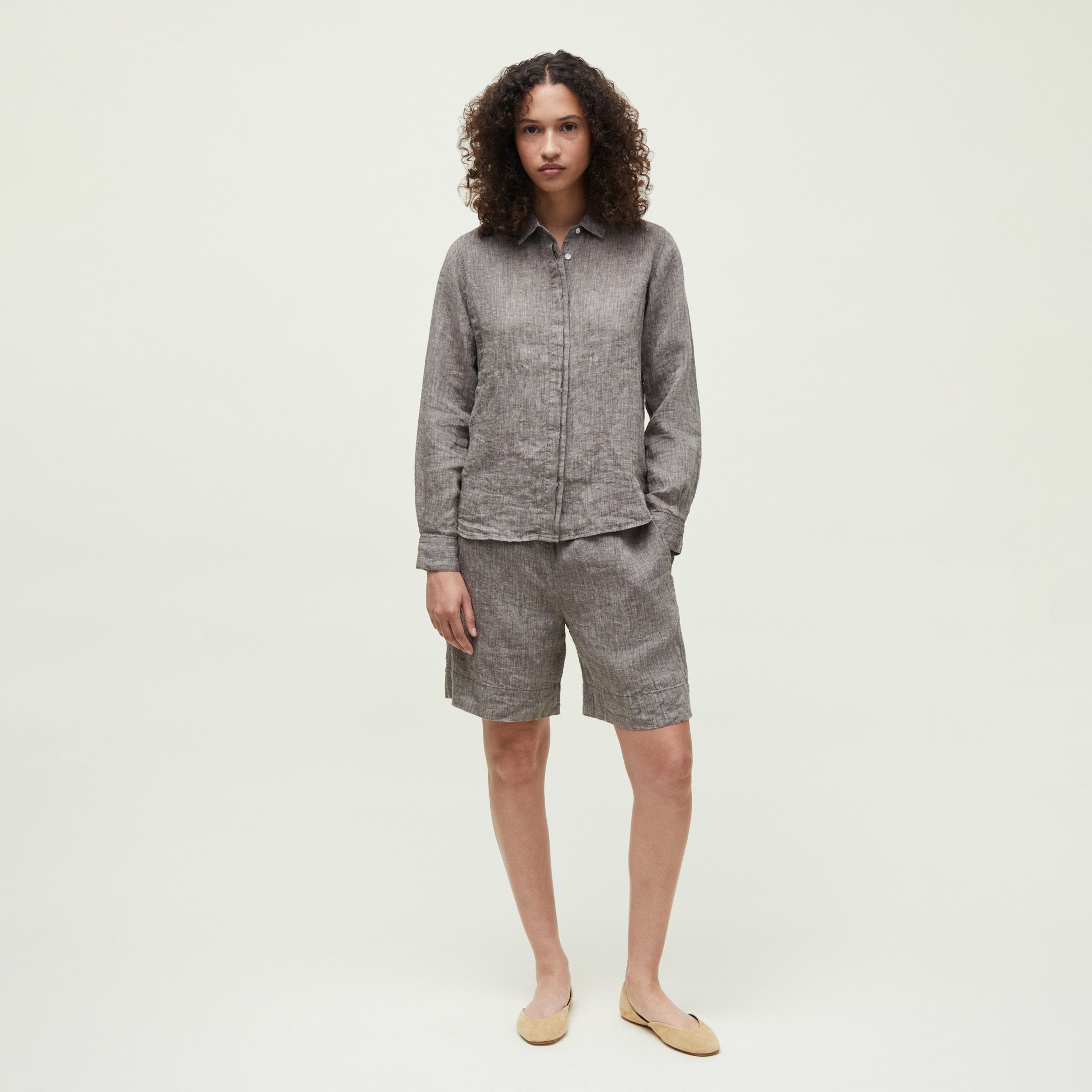 A person stands against a plain background wearing the Aiayu Carol Linen Shirt paired with matching knee-length shorts and flat shoes, creating a clean minimalist look.