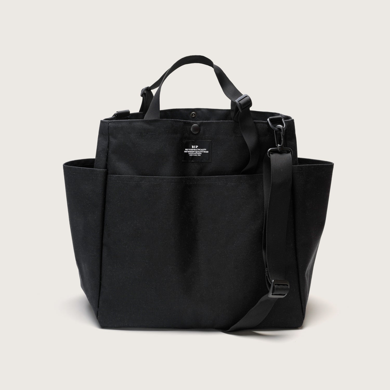 The Carry All Beach Bag by Bags in Progress is a black canvas and nylon tote with two short adjustable handles, a detachable shoulder strap, side pockets, and a small white front label, shown on a plain light background.