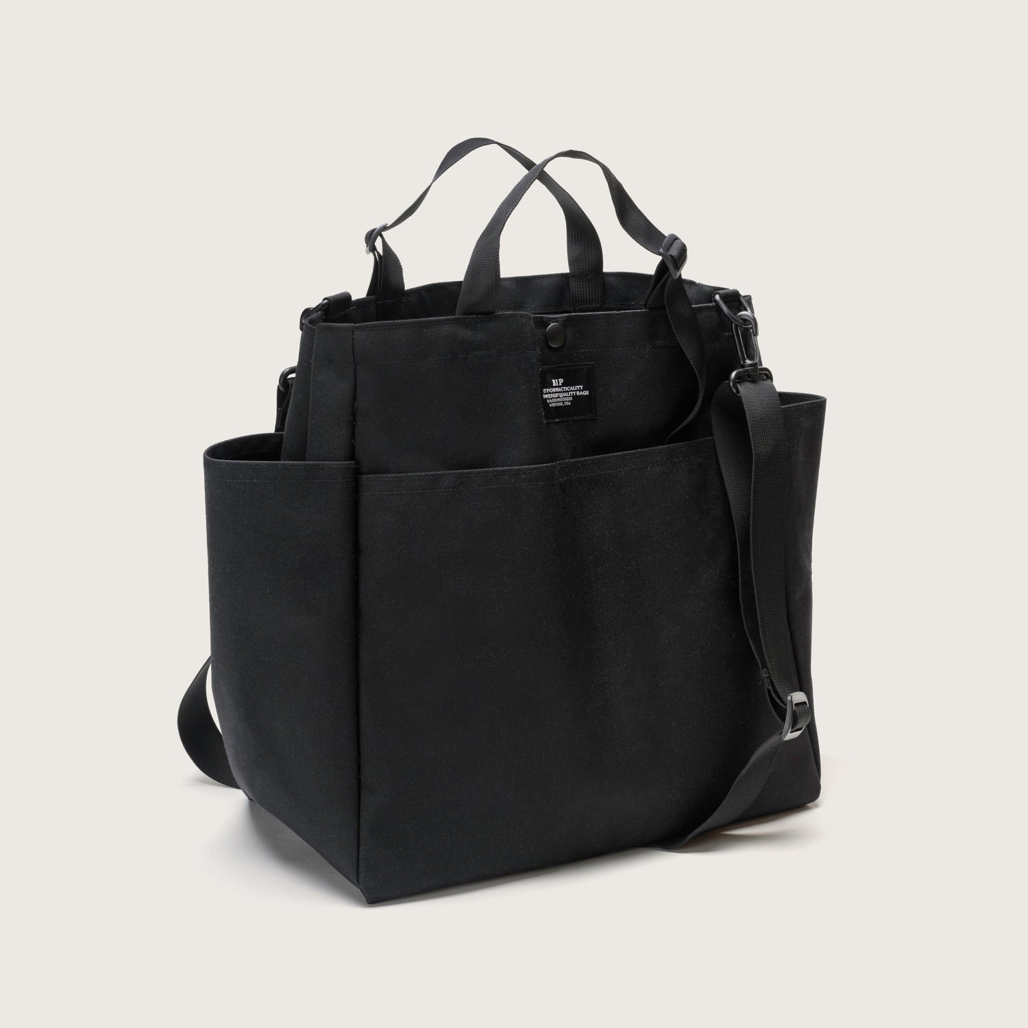The Carry All Beach Bag by Bags in Progress features black canvas, multiple exterior pockets, adjustable handles, a detachable shoulder strap, and a snap button closure, displayed against a plain light background.