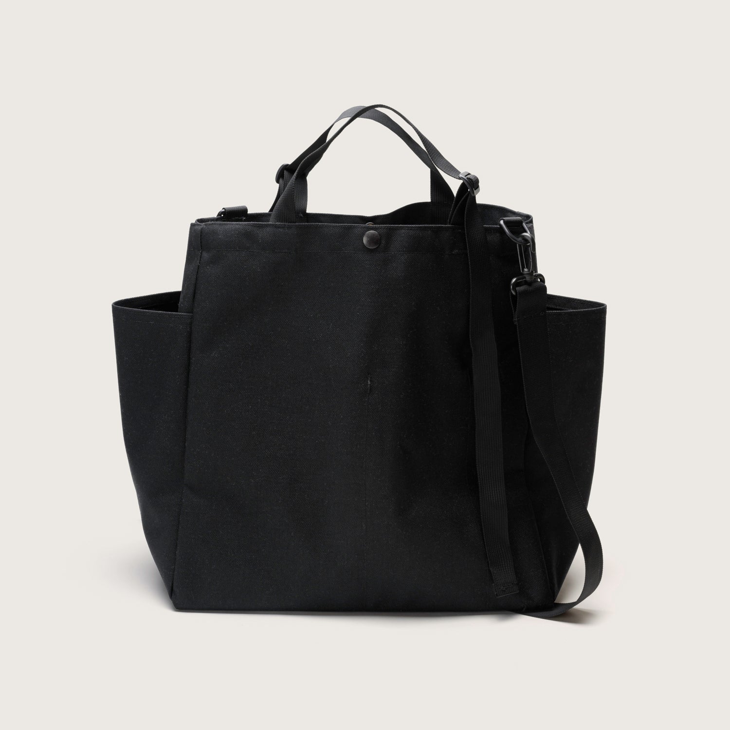 The Carry All Beach Bag by Bags in Progress is a black nylon tote with side pockets, short adjustable handles, and a detachable shoulder strap, shown against a plain light background.