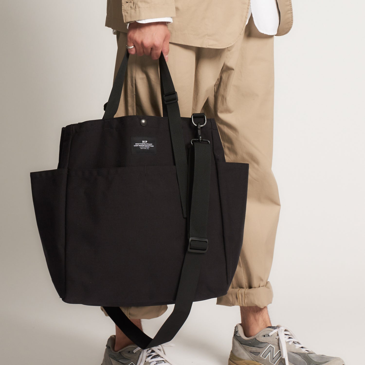 A person in tan pants and sneakers holds the Bags in Progress Carry All Beach Bag, a large black nylon tote with adjustable handles and a detachable shoulder strap, against a plain background.