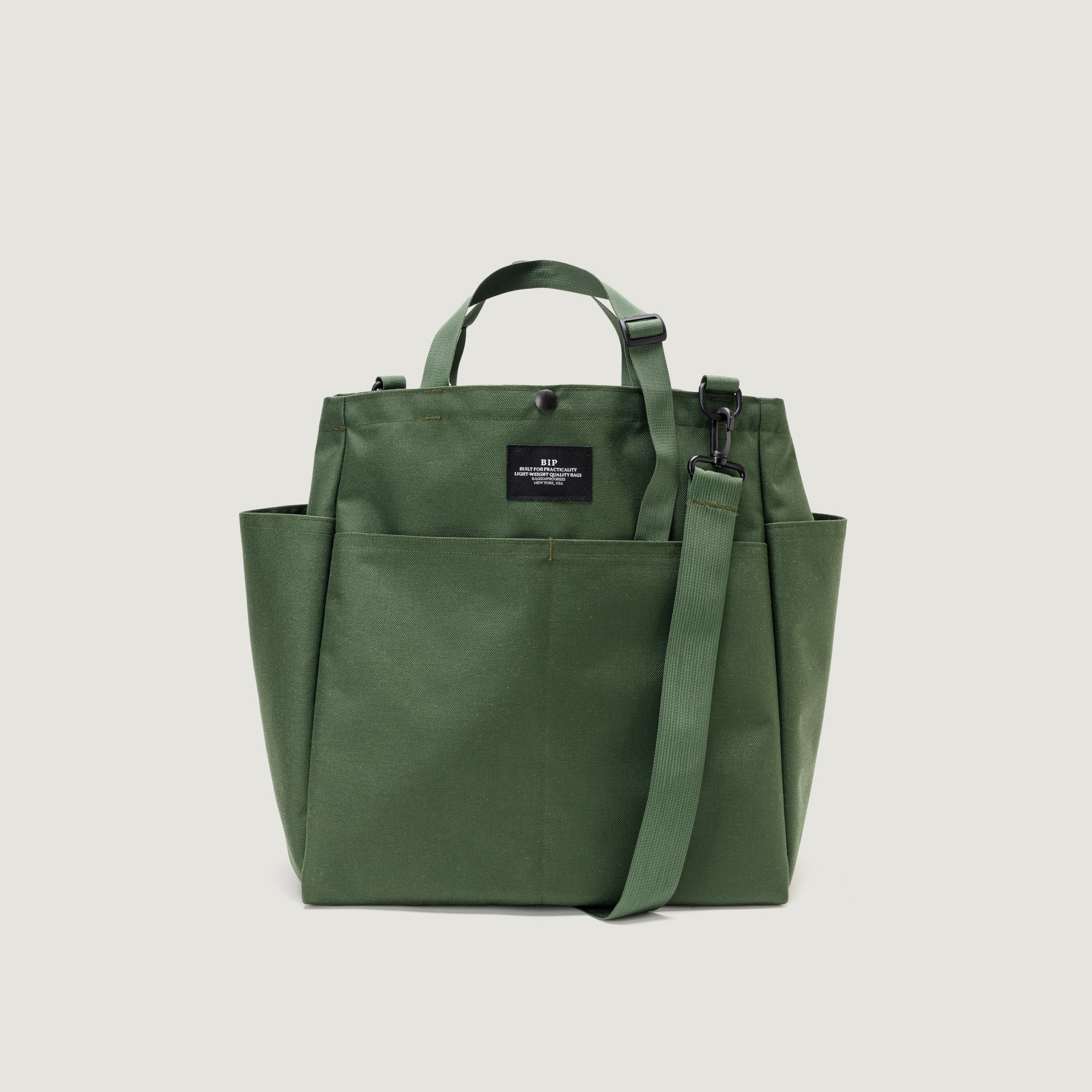 The Carry All Beach Bag by Bags in Progress is a green nylon tote with two handles, a detachable shoulder strap, side pockets, and a black front label, displayed on a plain white background.