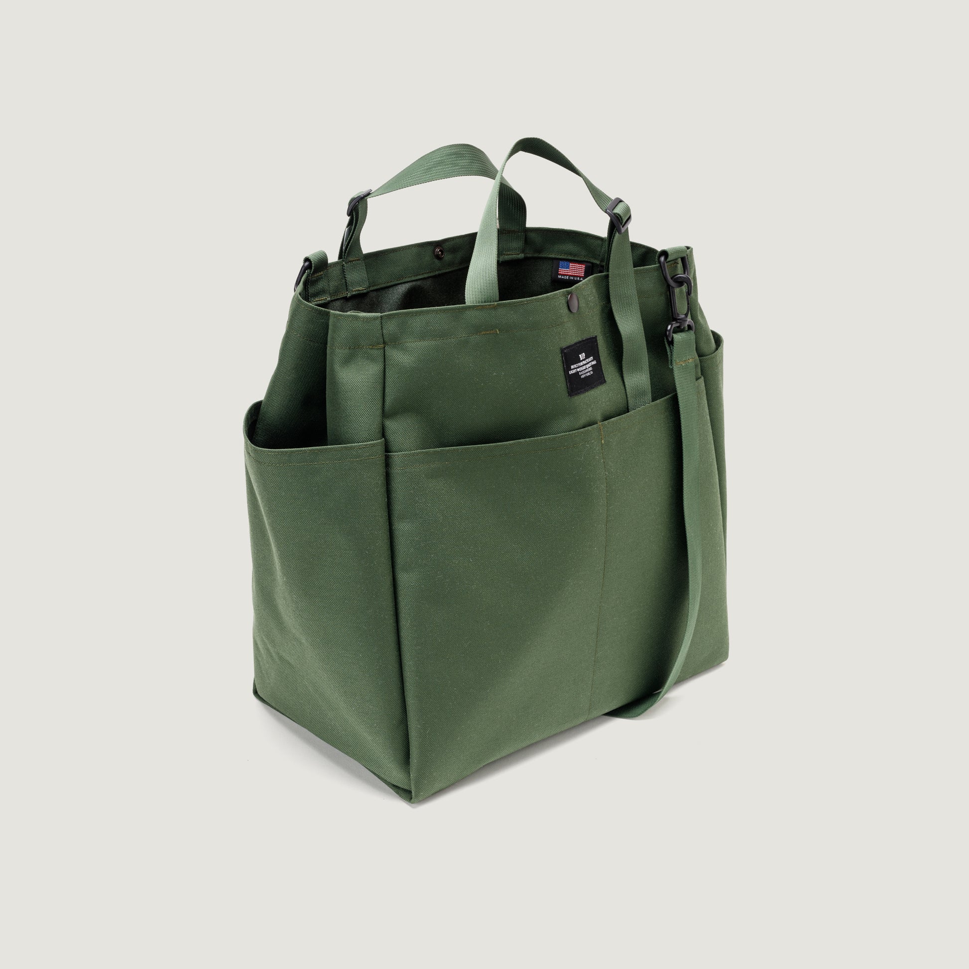 The Carry All Beach Bag by Bags in Progress is shown against a plain light background. This green canvas and nylon tote features adjustable handles, a detachable shoulder strap, exterior pockets, and a black label tag.