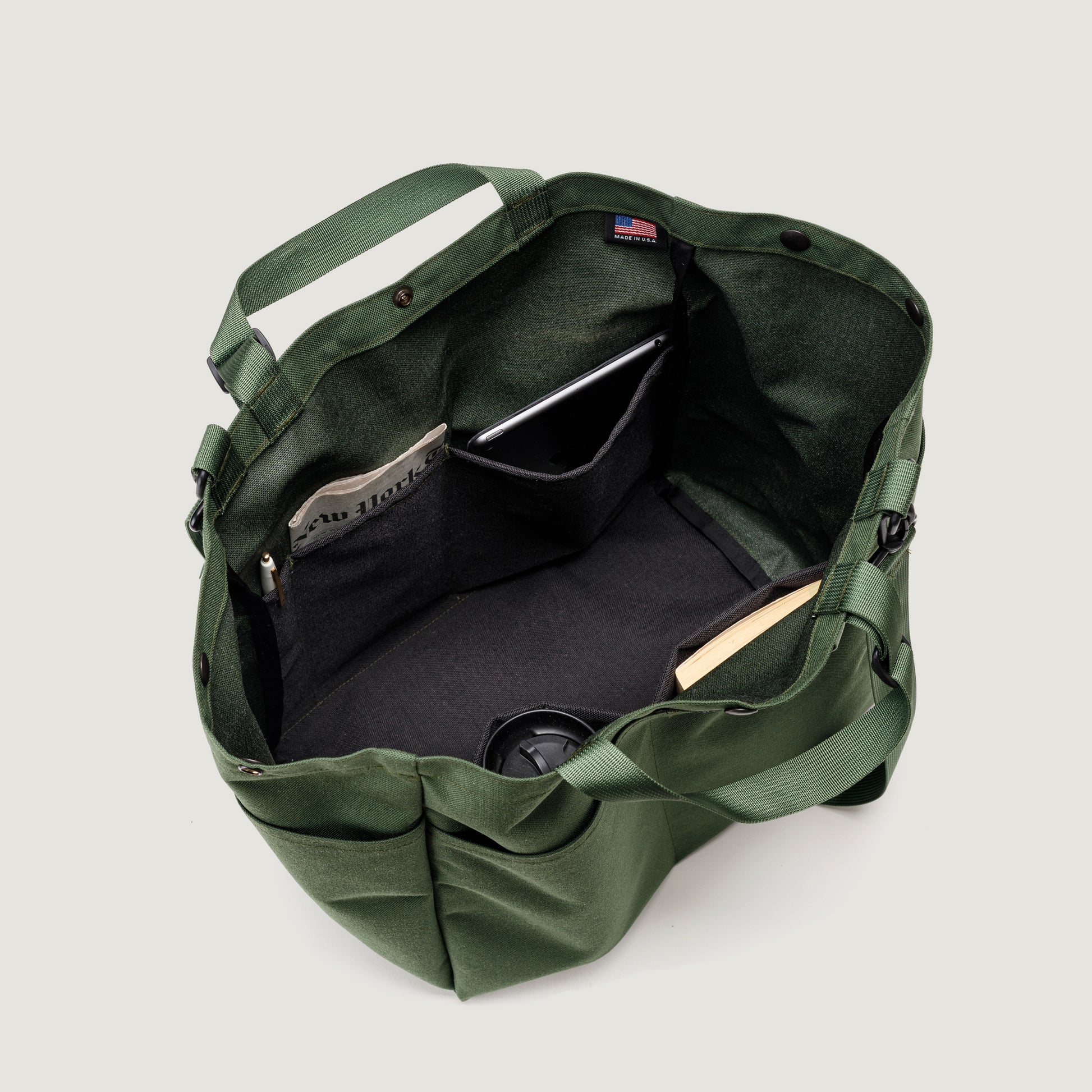 The Carry All Beach Bag by Bags in Progress is an open green nylon tote with adjustable handles and interior pockets holding a newspaper, tablet, book, and travel mug; photographed from above against a plain background.