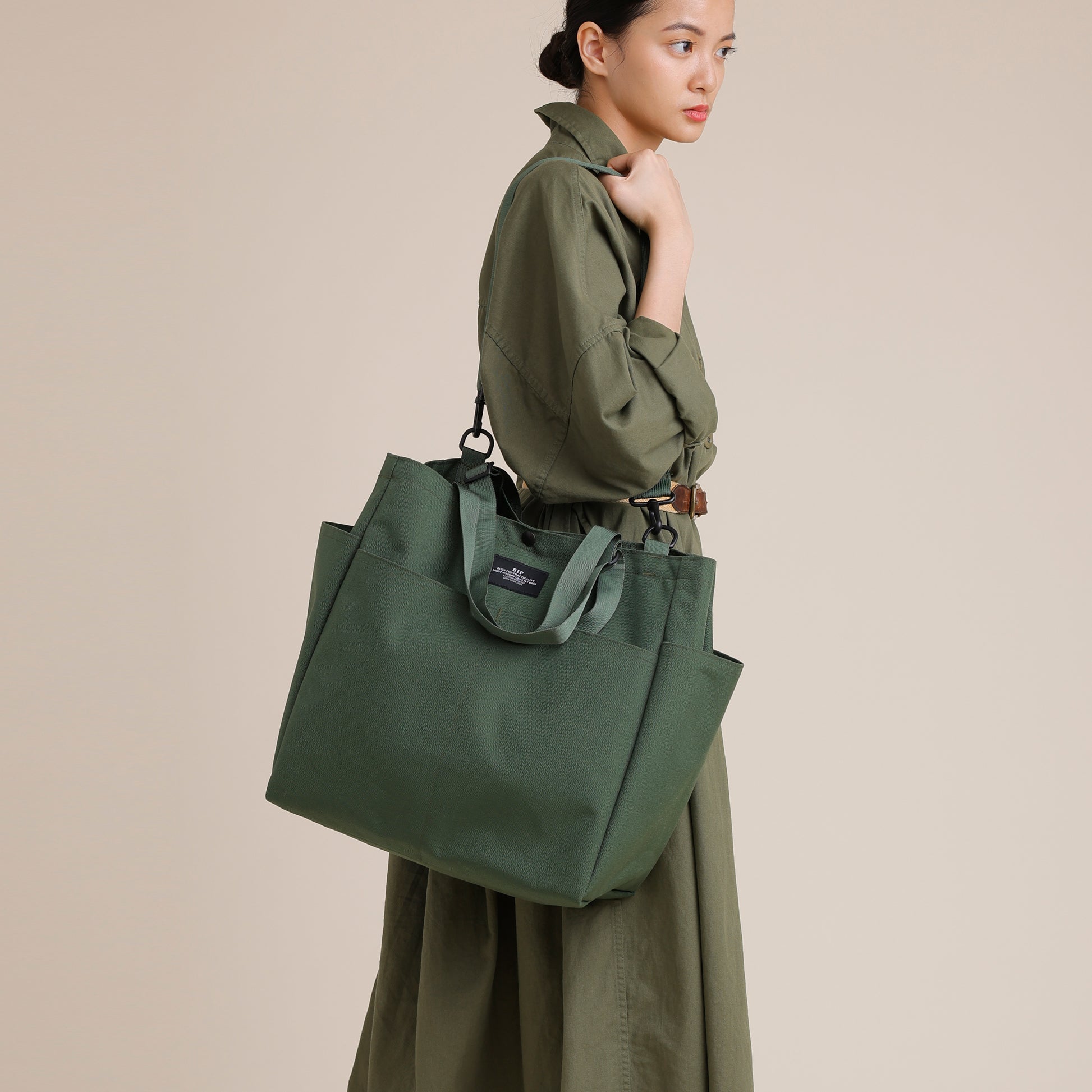 A woman in a long-sleeved green dress carries the Bags in Progress Carry All Beach Bag, a spacious nylon tote with adjustable handles, over her shoulder, standing against a plain beige background.
