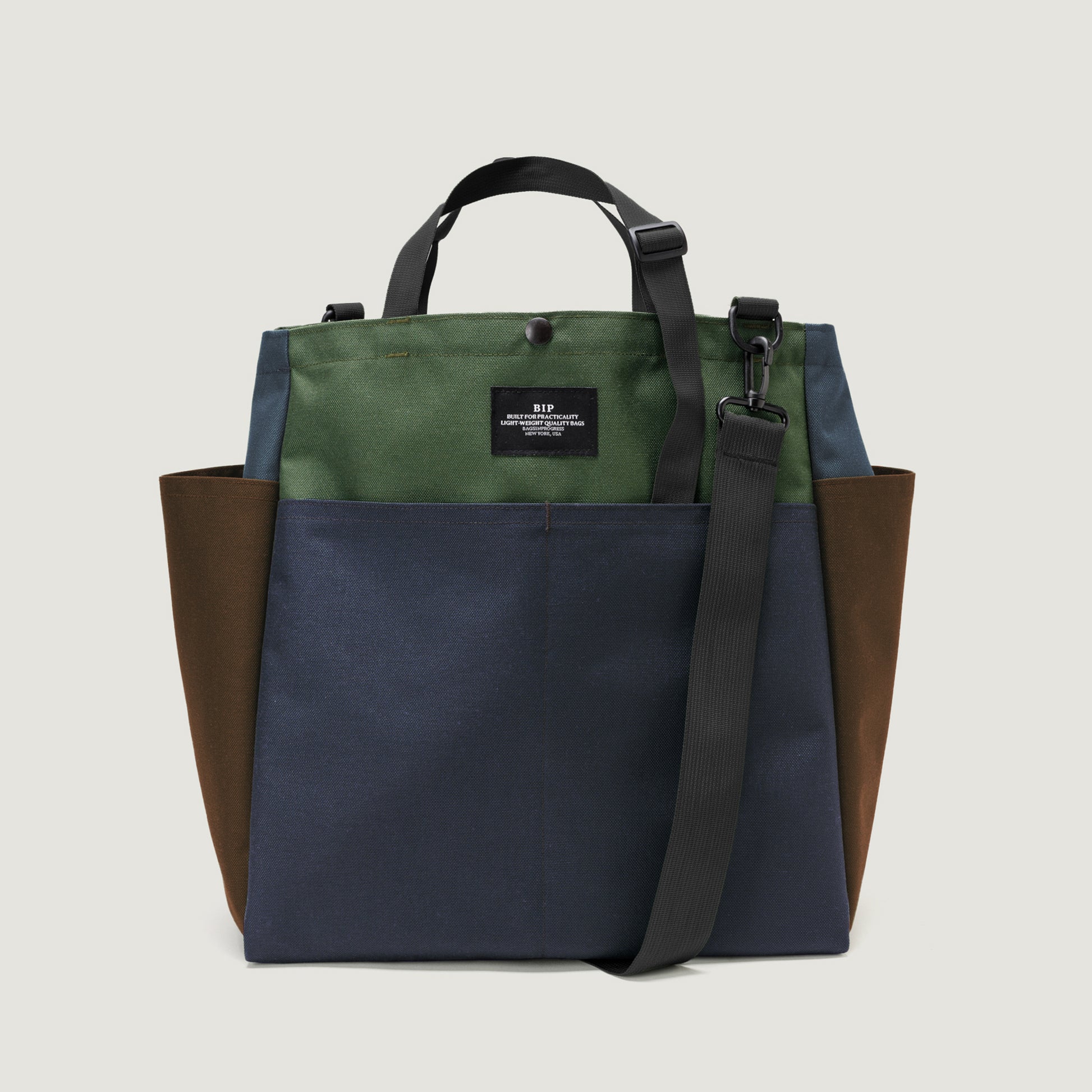 The Carry All Beach Bag by Bags in Progress is a color-blocked nylon tote with green, blue, and brown panels, black handles, an adjustable/detachable shoulder strap, and side pockets, shown against a light background.