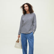 A person stands against a plain light background wearing Aiayu’s Circular Long Sleeve II—a black and white striped dropped-shoulder top—with blue jeans and holding a beige handbag.