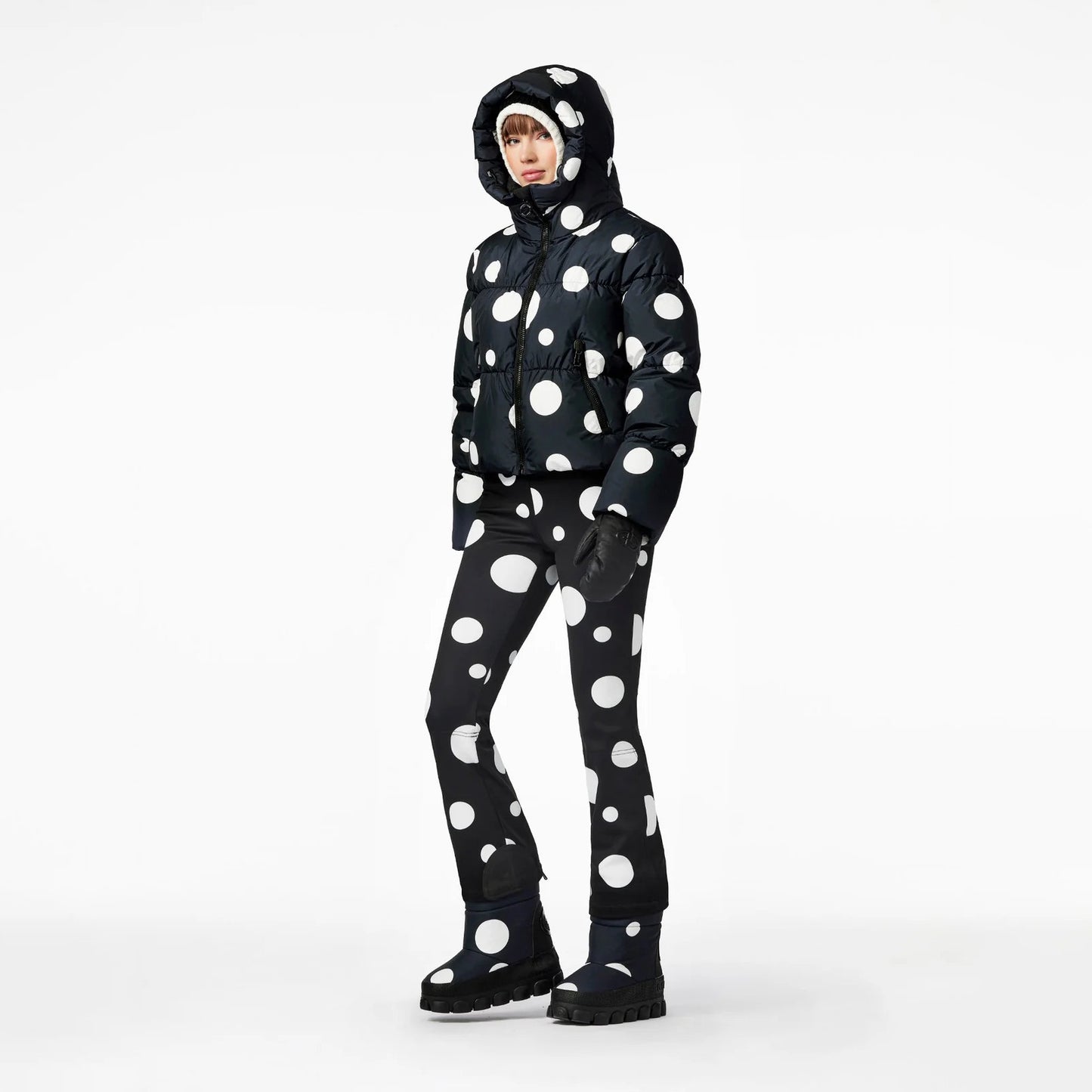 A person stands against a plain background wearing Goldbergh's Grand Ski Jacket in Dot Black, featuring large white polka dots, with matching pants and boots.