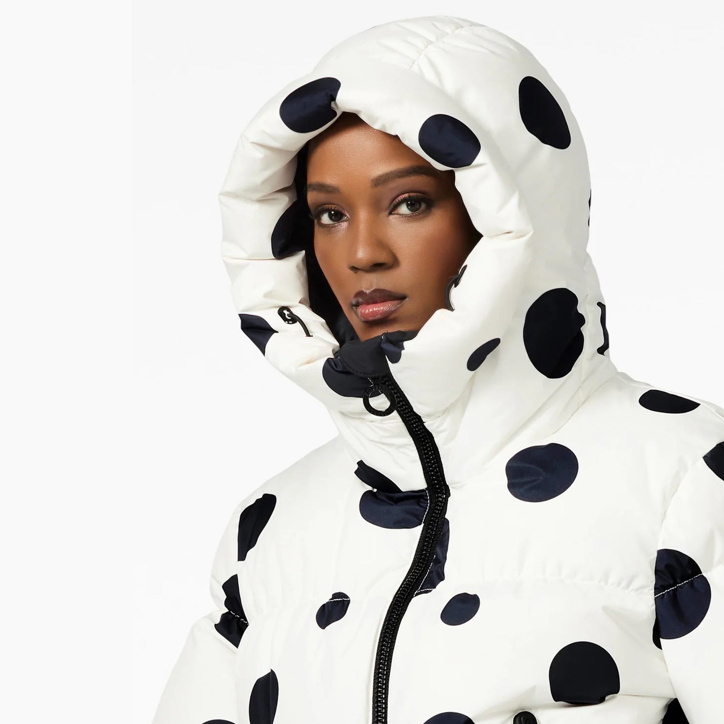 A person wears the Grand Ski Jacket, Dot Cream by Goldbergh. The hood is up, and the jacket is zipped tight, providing excellent insulation. Its bold design features large black polka dots on white—contrasting beautifully against a crisp background with a neutral expression.