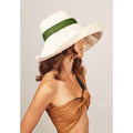 A woman with wavy hair wears a large, white sunhat reminiscent of sail canvas with a green band and a tan, one-shoulder top, facing to the side against a plain background for optimal sun protection. She is wearing the Dia, Natural / Grass by Lola Hats.