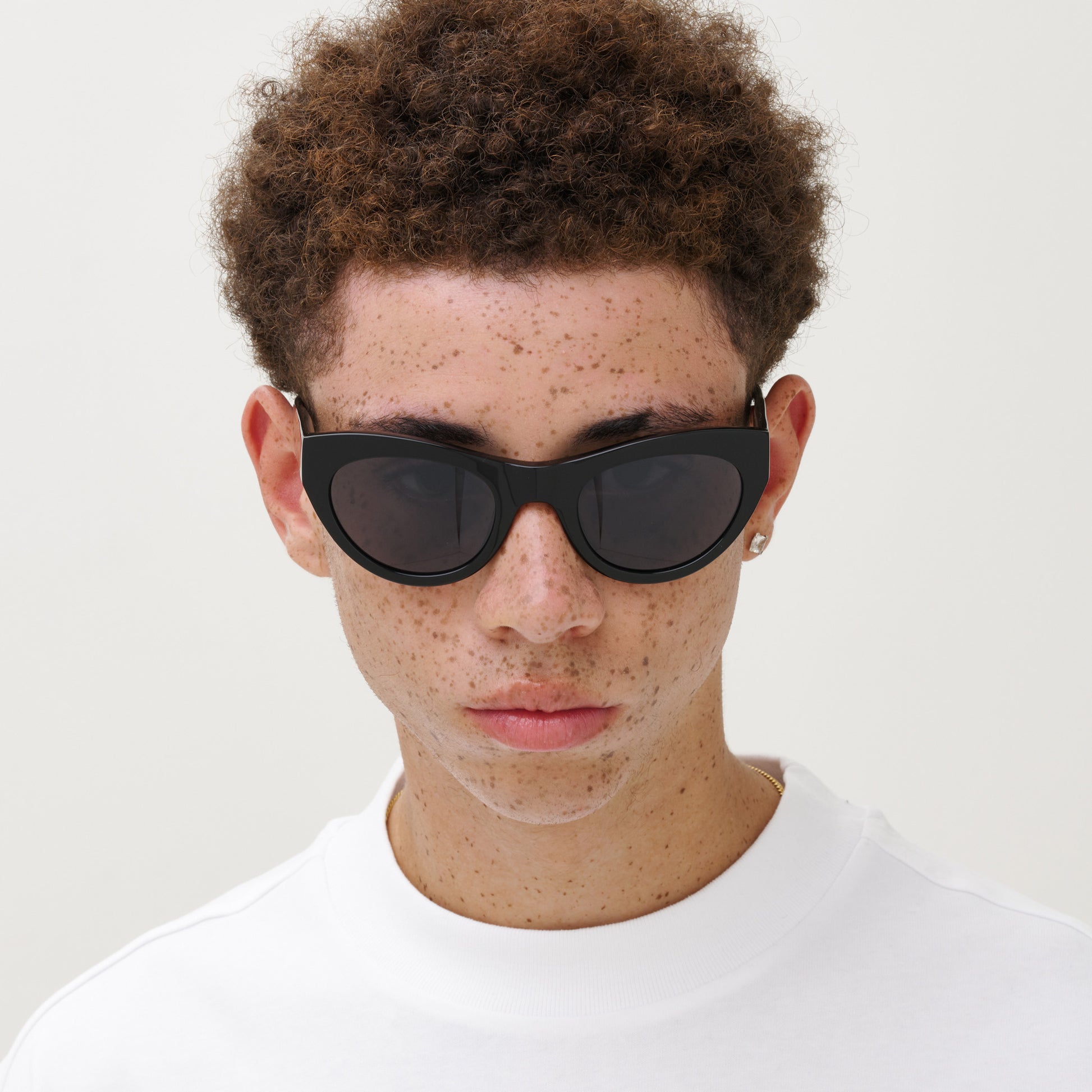 A person with short curly hair and freckles wears Messyweekend’s Donna sunglasses in Grey/Black and a white top, facing the camera against a plain, light background.