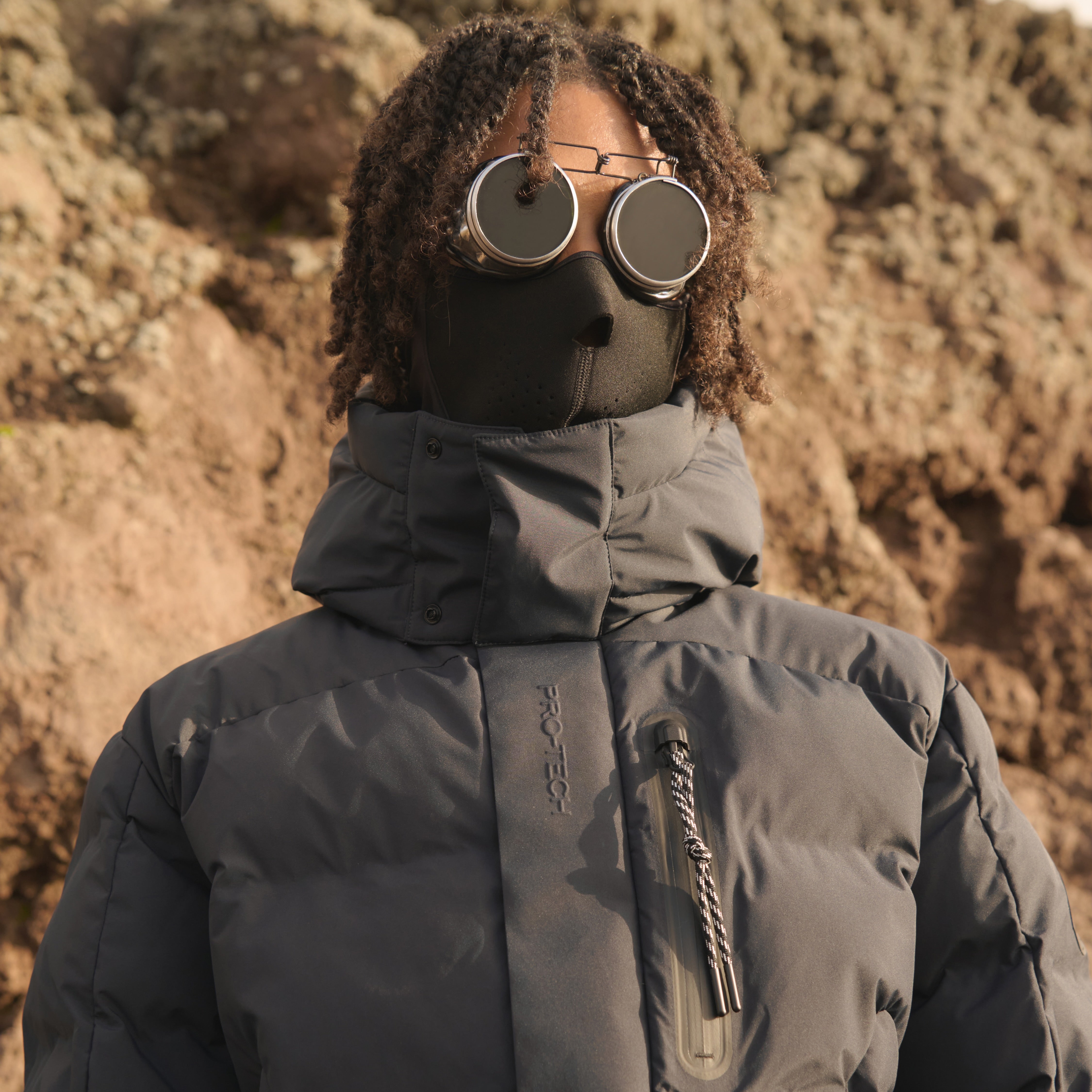 Person wearing a black jacket and goggles in front of a rocky background