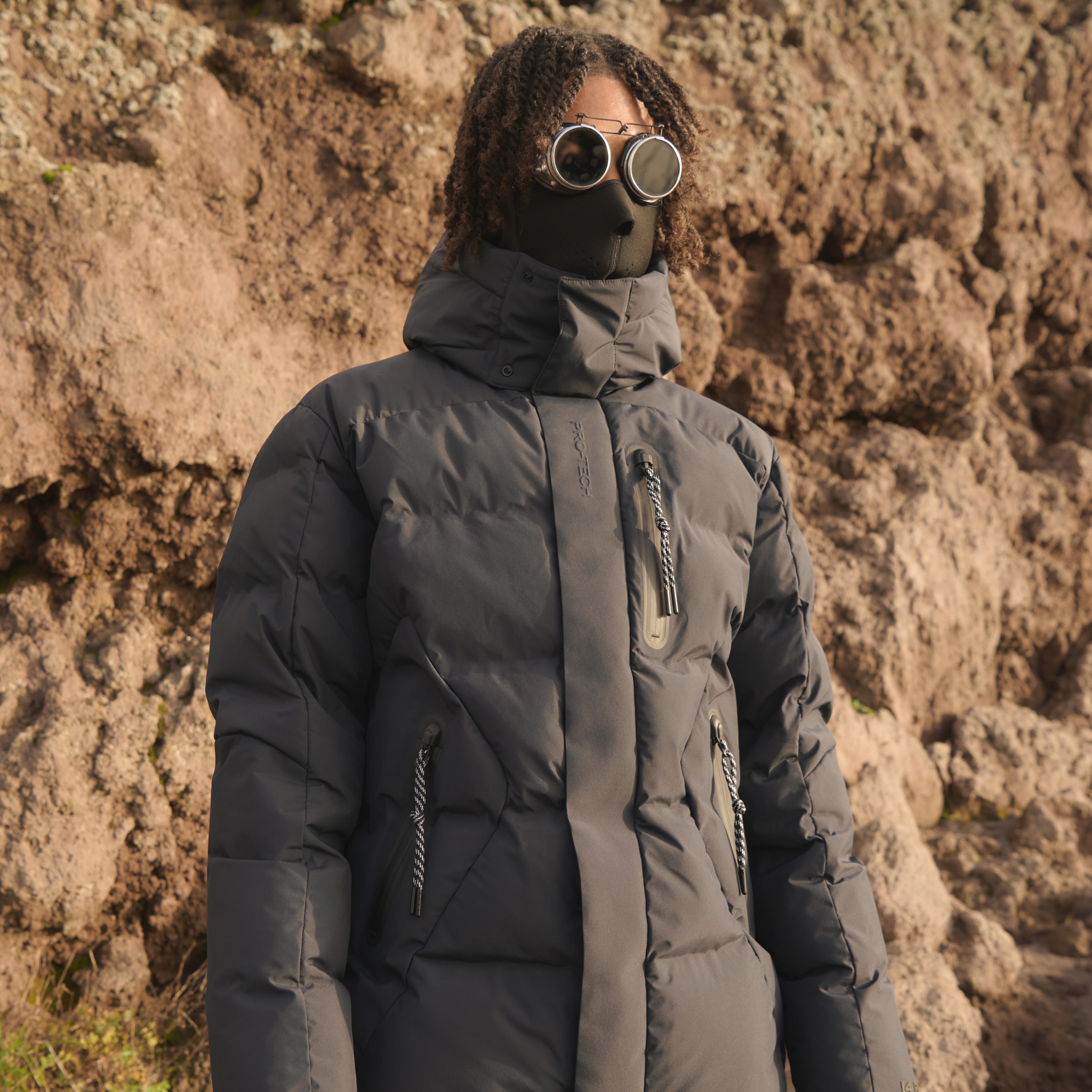 Person wearing a dark gray puffer jacket and goggles against a rocky background