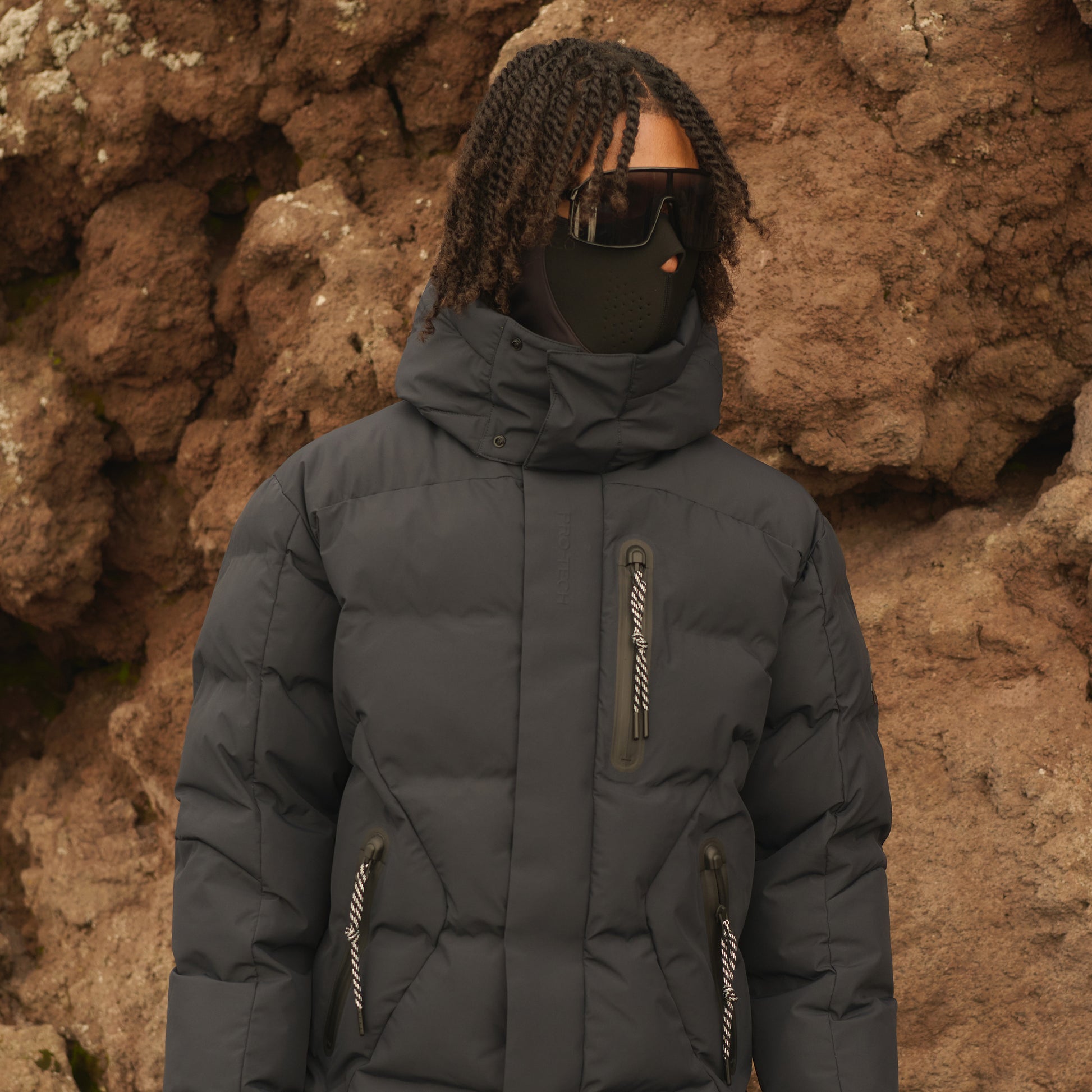 Person wearing a dark gray puffer jacket and sunglasses against a textured brown wall.