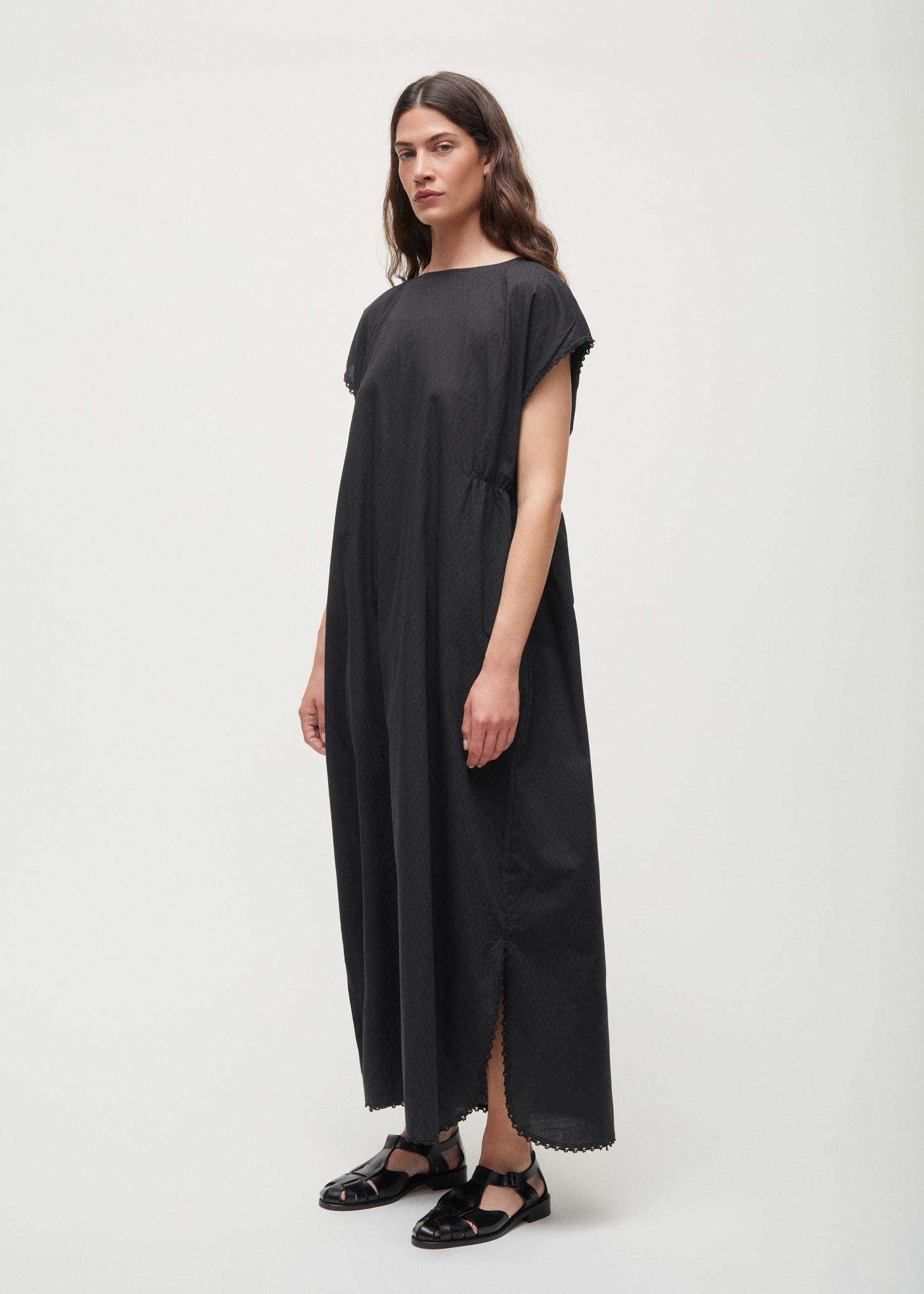Against a plain backdrop, a person dons the Aiayu Diamond Dress, a sleeveless, loose-fitting maxi crafted from GOTS-certified cotton, paired with black sandals.