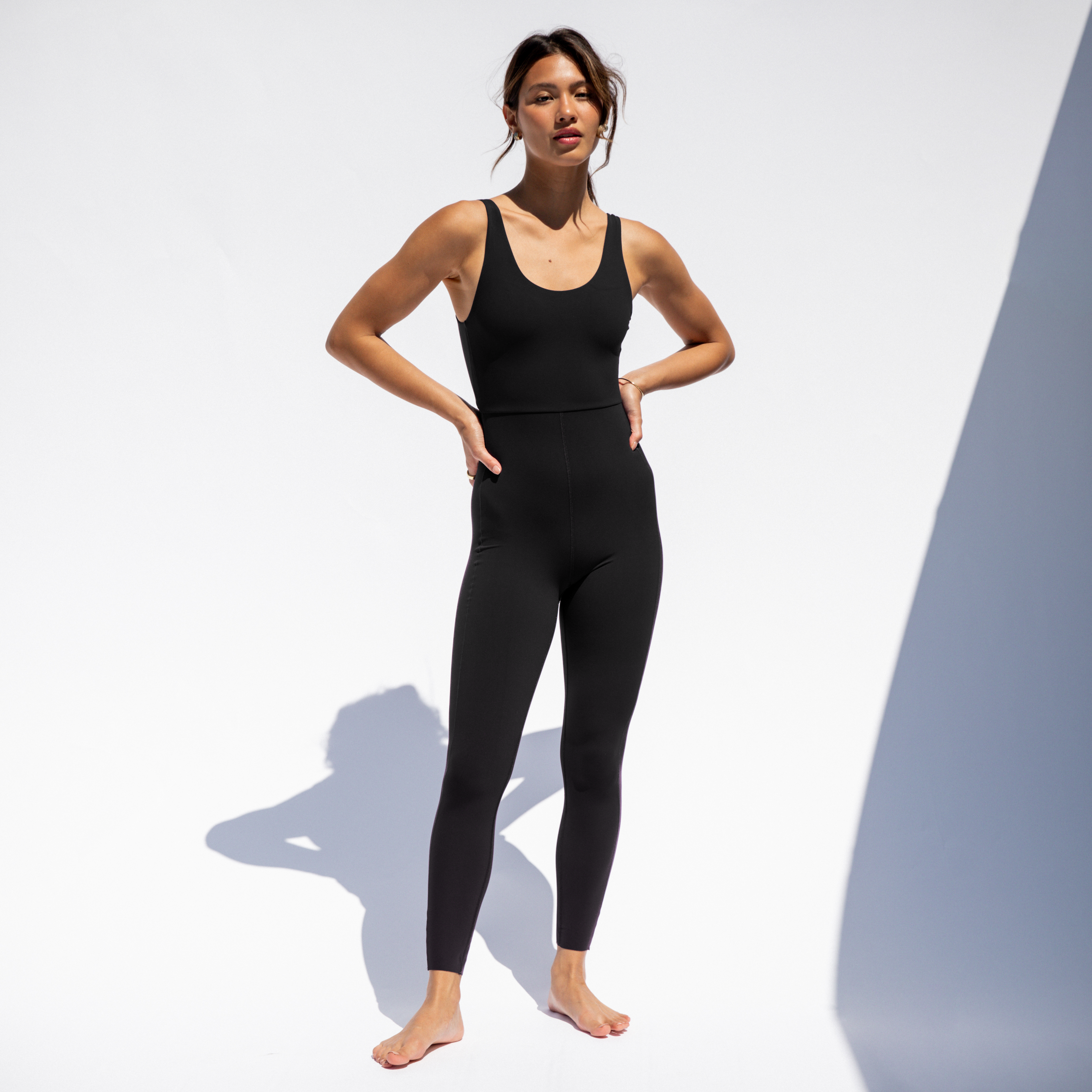 A person stands barefoot against a white background, wearing the Double Scoop Playsuit 27" by Left On Friday—a fitted black sleeveless jumpsuit with an ultra-comfortable fit, perfect for low-impact activities and posing hands on hips.