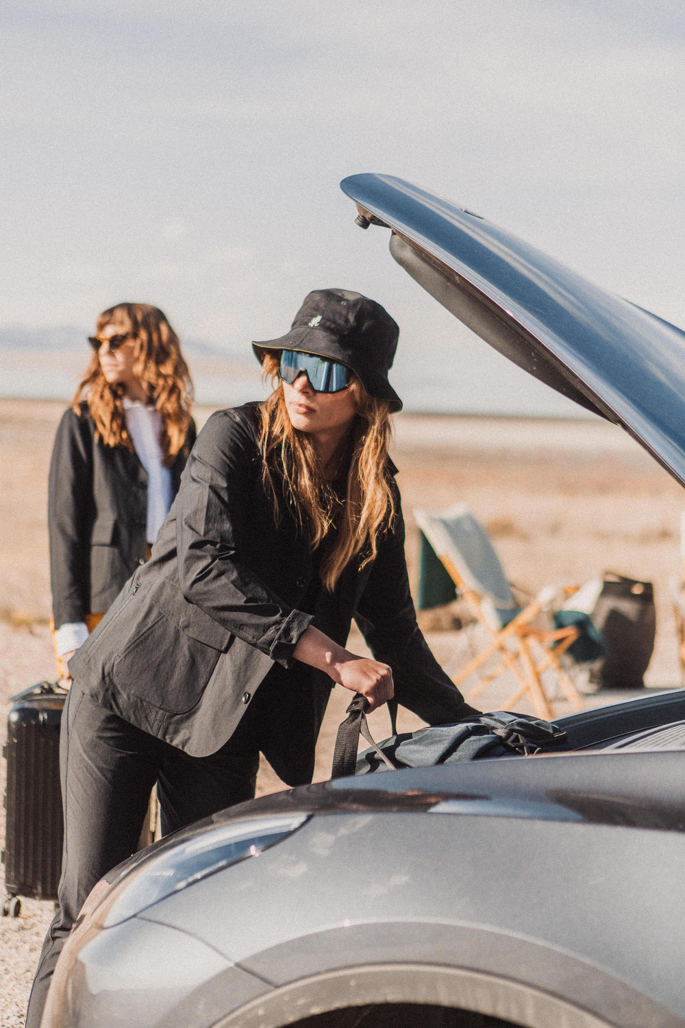 female model wearing dark suit, bucket hat and sunglasses unloading trunk, backround in blur camping chair and another female