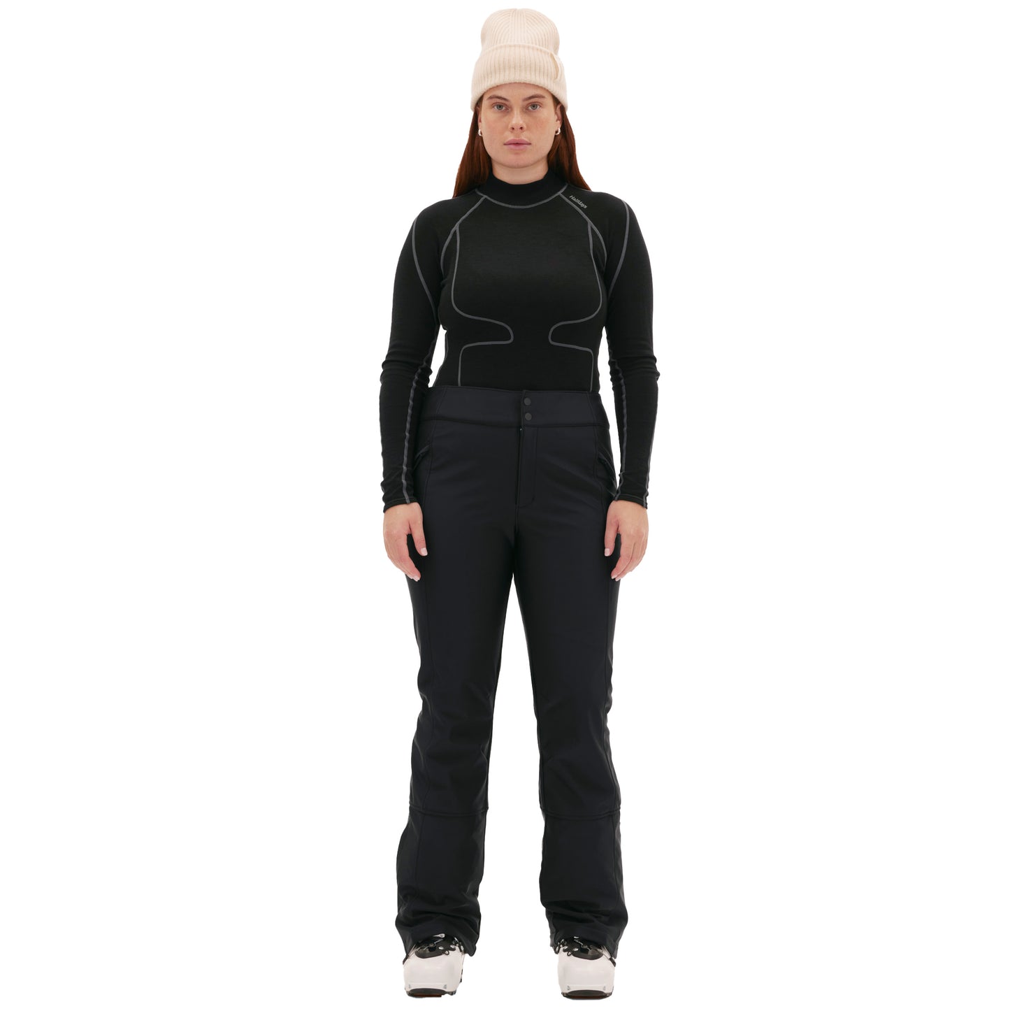 Against a white background, a person wears a black long-sleeve top, Halfdays Emma Pant ski pants in a slim fit, ski boots, and a white beanie.
