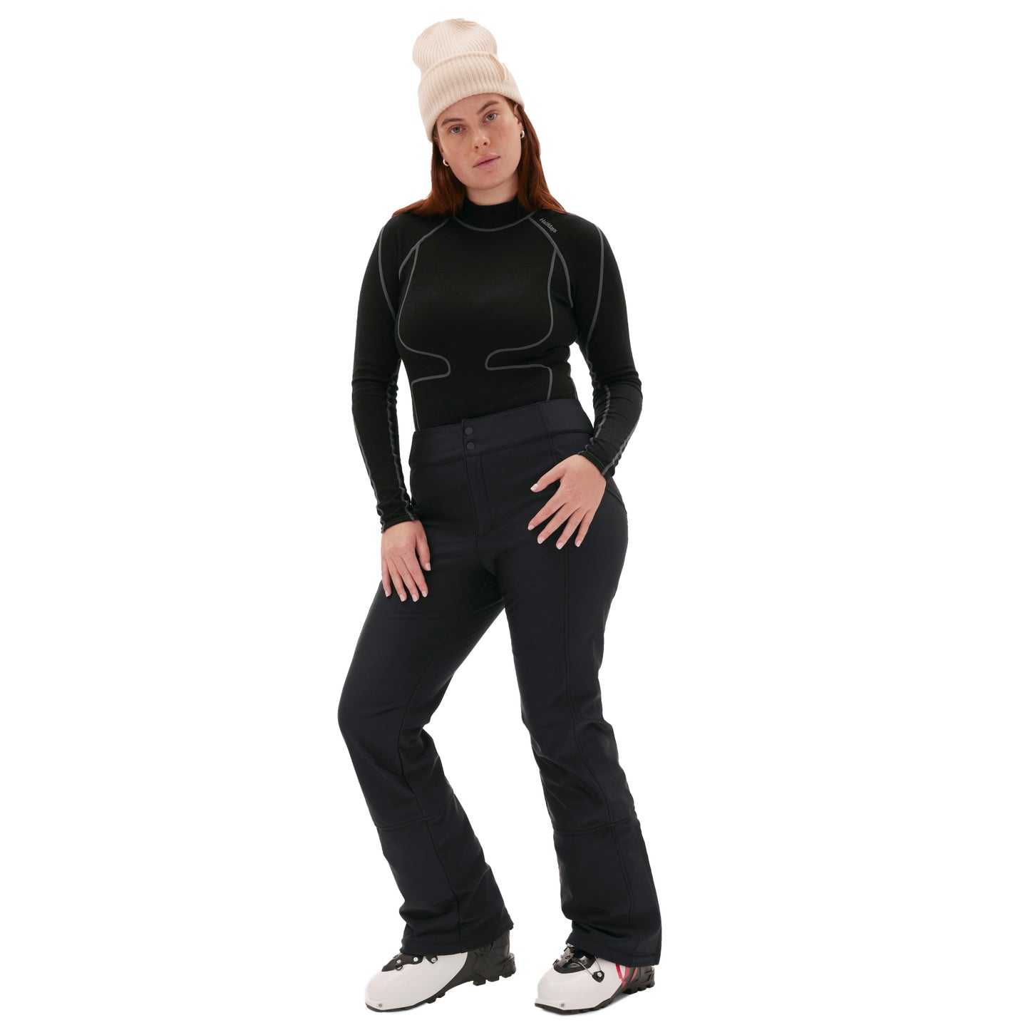 A person in a black outfit and beige beanie stands with one hand on their hip, wearing the Emma Pant from Halfdays and white boots, creating a sleek silhouette against the plain background.