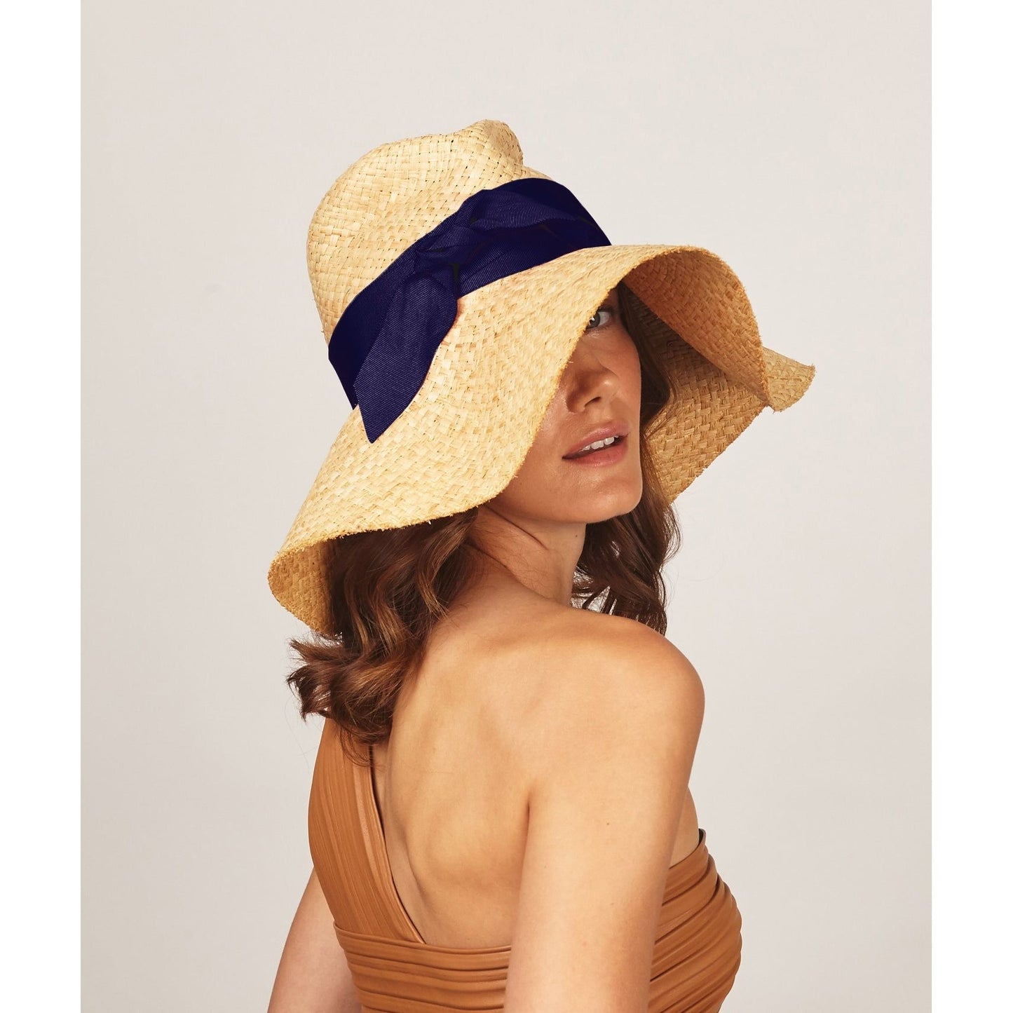 A woman with wavy brown hair wears a wide-brimmed raffia material straw hat called "First Aid, Nude w/ Navy" by Lola Hats, a one-shoulder brown top, and looks over her shoulder against a neutral background.