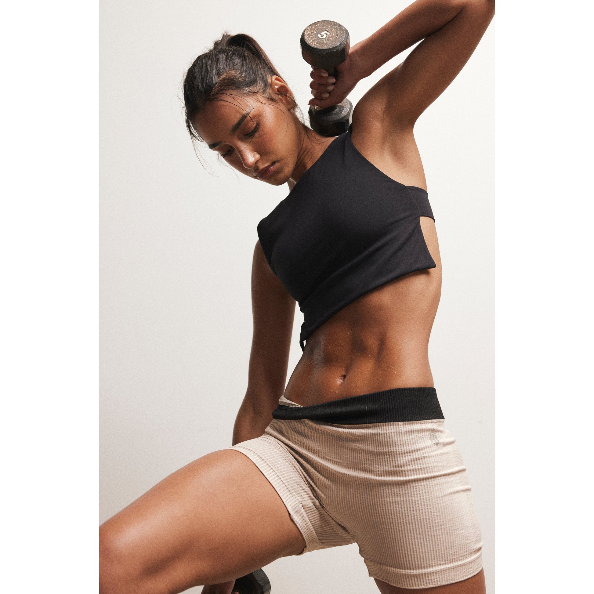 A fit woman lifting a dumbbell, focusing intensely, showcasing toned abdominal muscles, wearing the Free People Movement Be Right Back Cami in black and beige shorts.