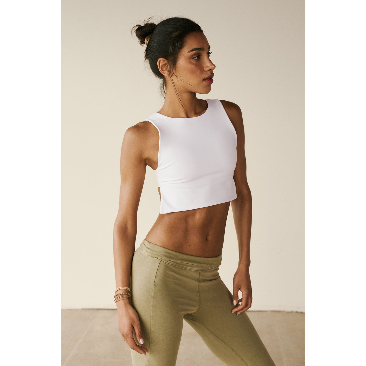A woman in a Free People Movement white Be Right Back Cami crop top and beige leggings stands in a neutral-toned room, looking to the side with a relaxed posture.