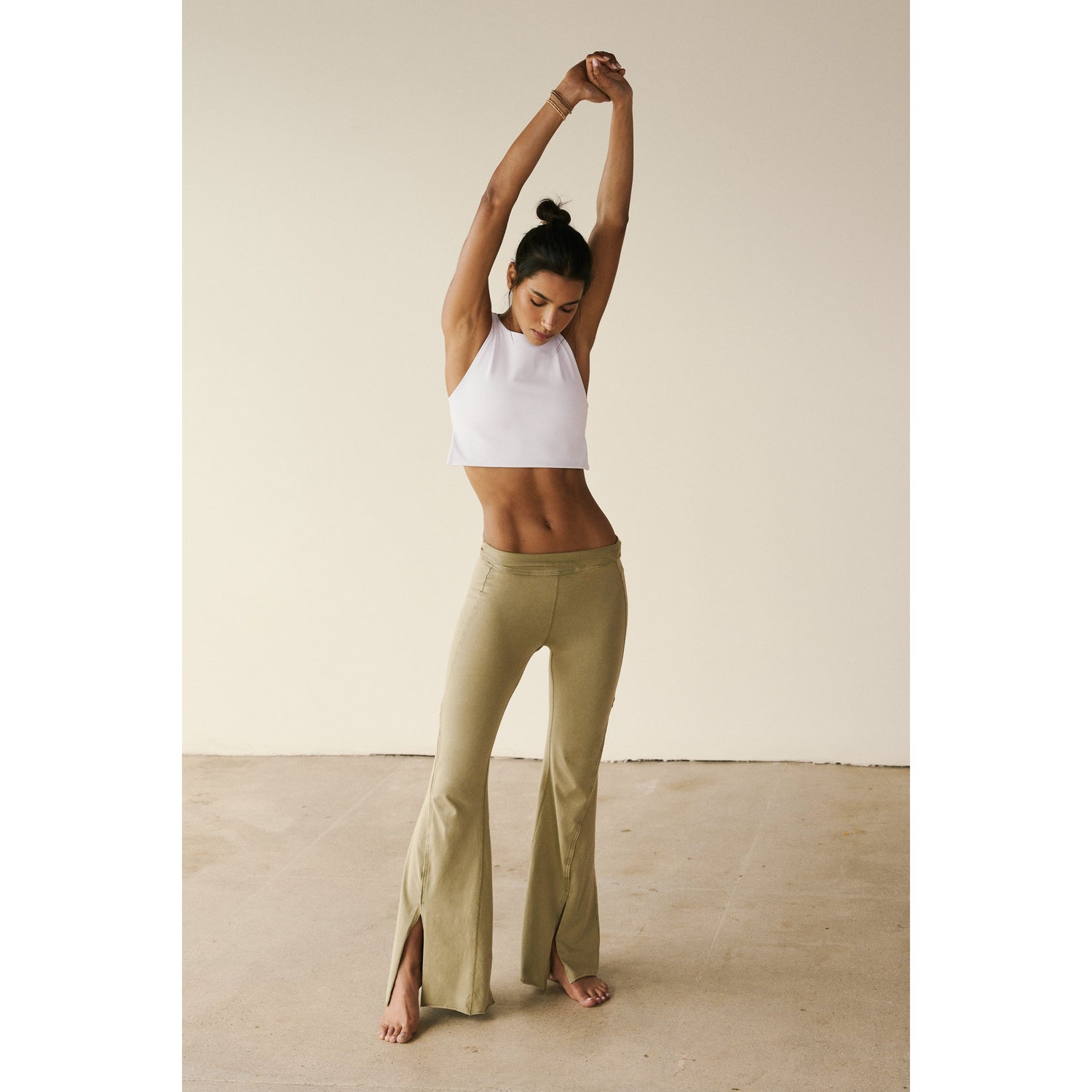 A woman in a Free People Movement Be Right Back Cami in White and green pants stretches her arms above her head in a bright room.