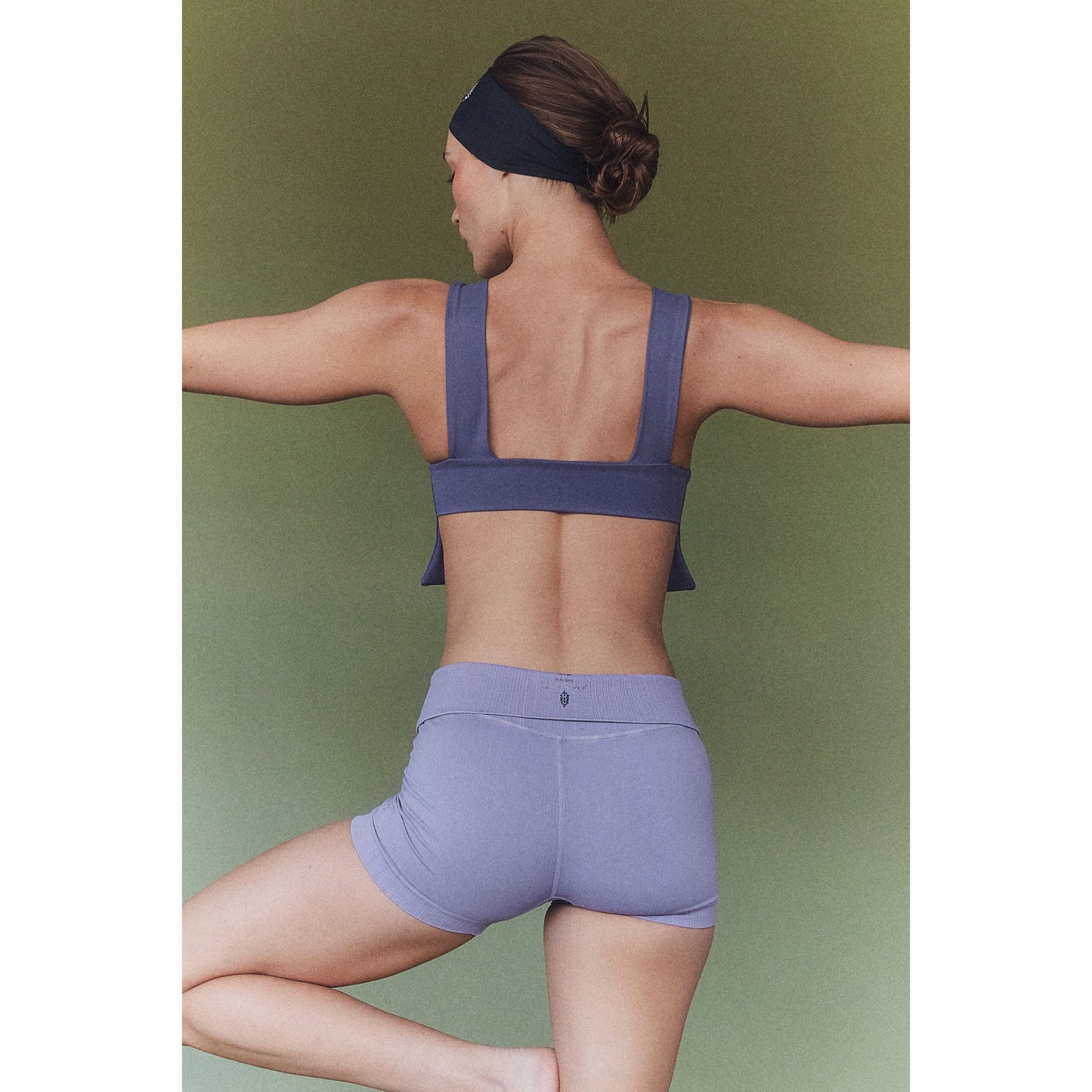 A woman in a purple Be Right Back Cami sports bra and shorts made of seamless fabric, stretching her arms back, wearing a headband, against a green background.