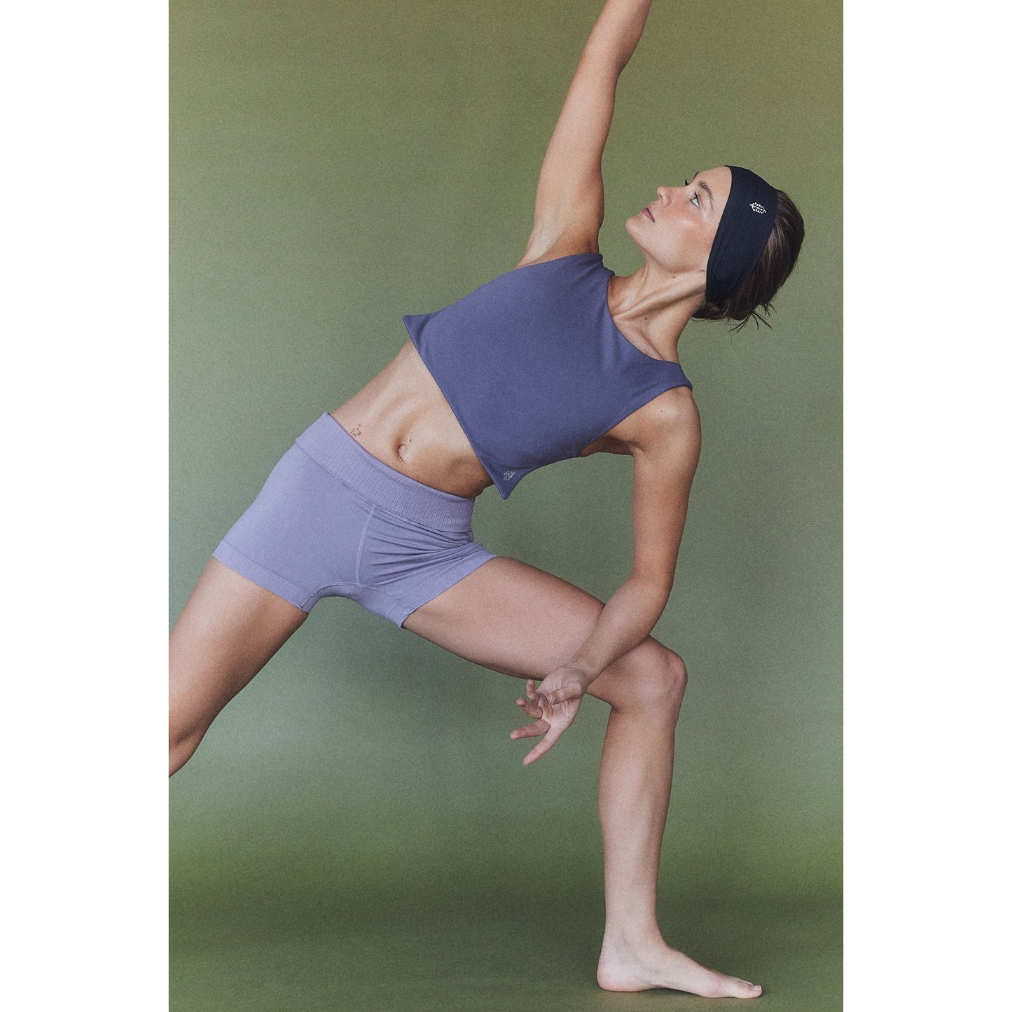 A woman in a Free People Movement Be Right Back Cami in Purple and shorts, featuring a strappy open back, performs a dynamic yoga pose against a green background.