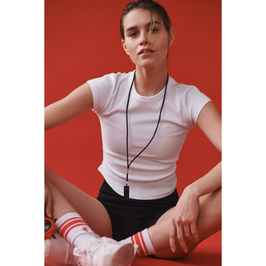 A young woman in sporty attire sits against a red background, wearing a Ribbed Baby Tee in White by Free People Movement, black shorts, striped socks, and sneakers, with a whistle around her neck.