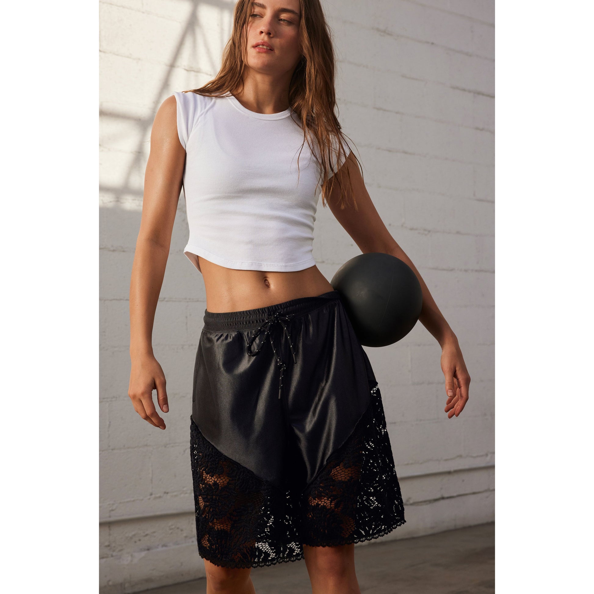 Woman in a Ribbed Baby Tee in White and black lace-trimmed shorts holding a medicine ball against a white wall by Free People Movement.