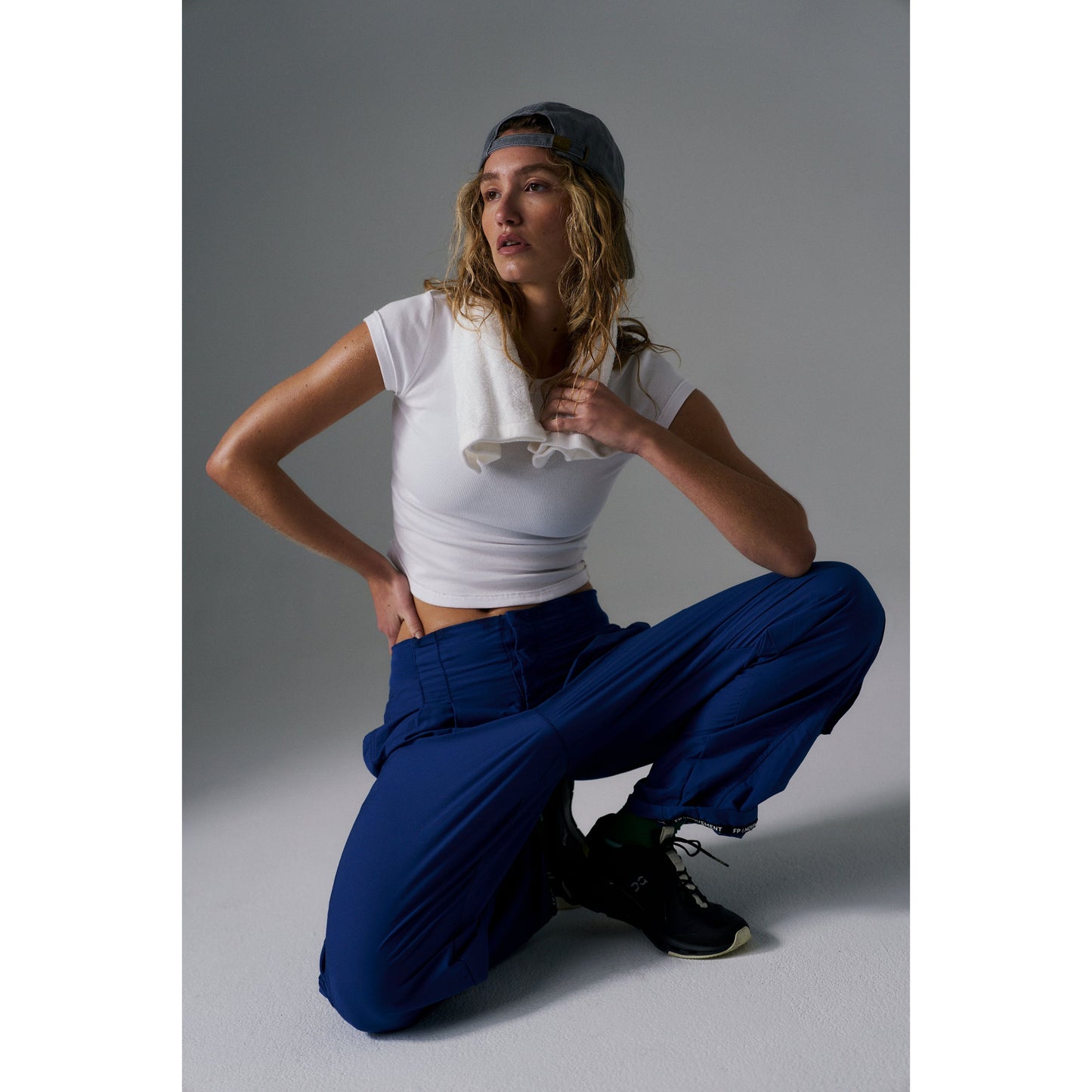 Woman in Free People Movement's Ribbed Baby Tee in White and blue pants, wearing a gray cap, sitting and looking to the side against a gray background.