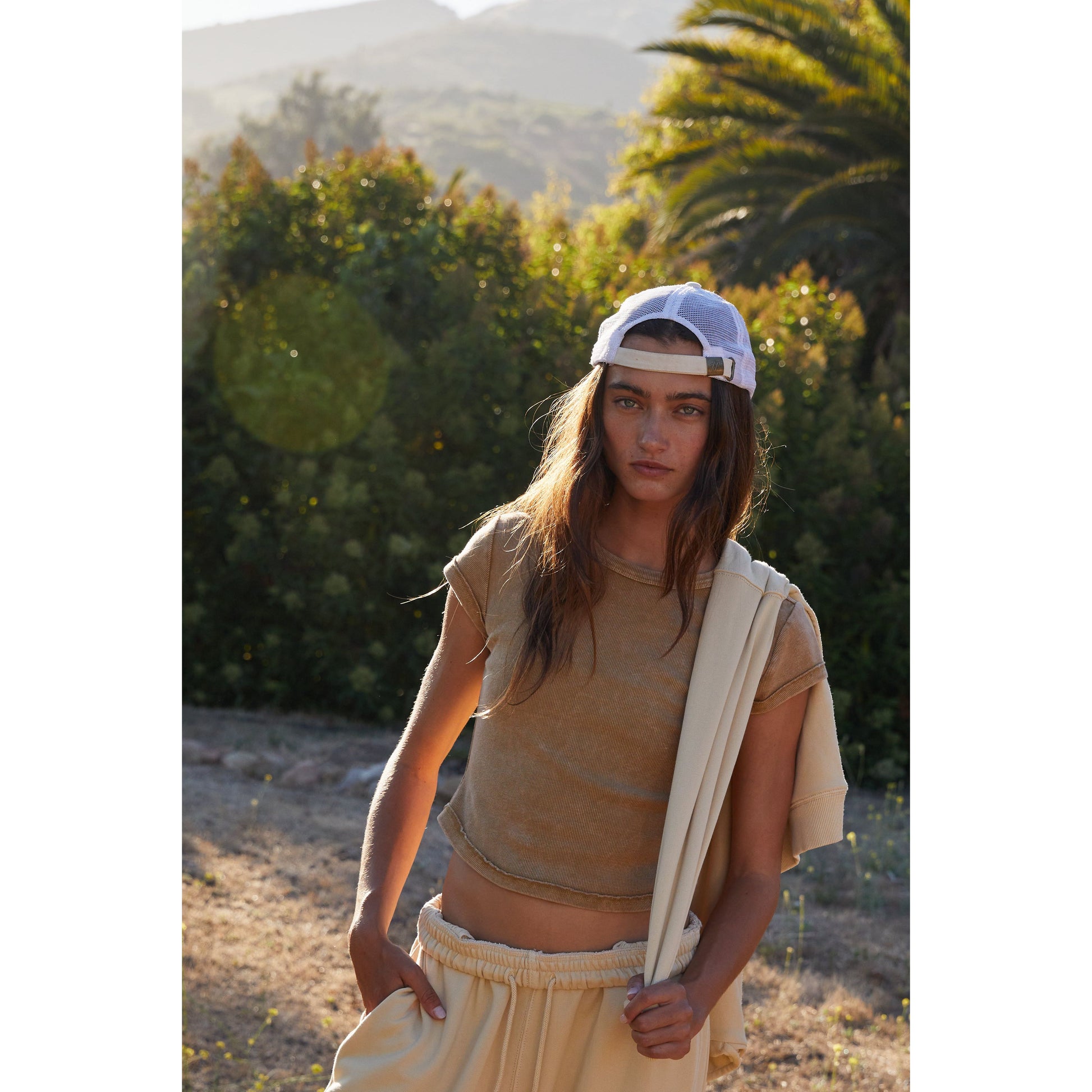 Young woman with a white cap and Antelope Ribbed Baby Tee carrying a canvas, standing in front of a sunlit hill with palm trees from Free People Movement.