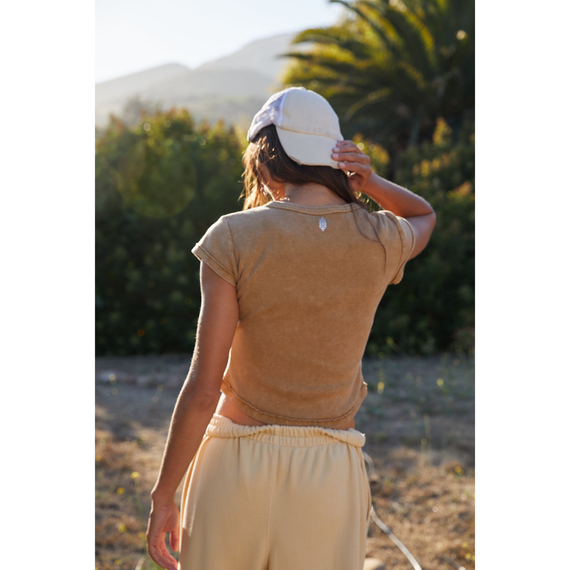 A woman in a beige Ribbed Baby Tee by Free People Movement and white hat stands outdoors, facing away from the camera, with mountains and palms in the background.