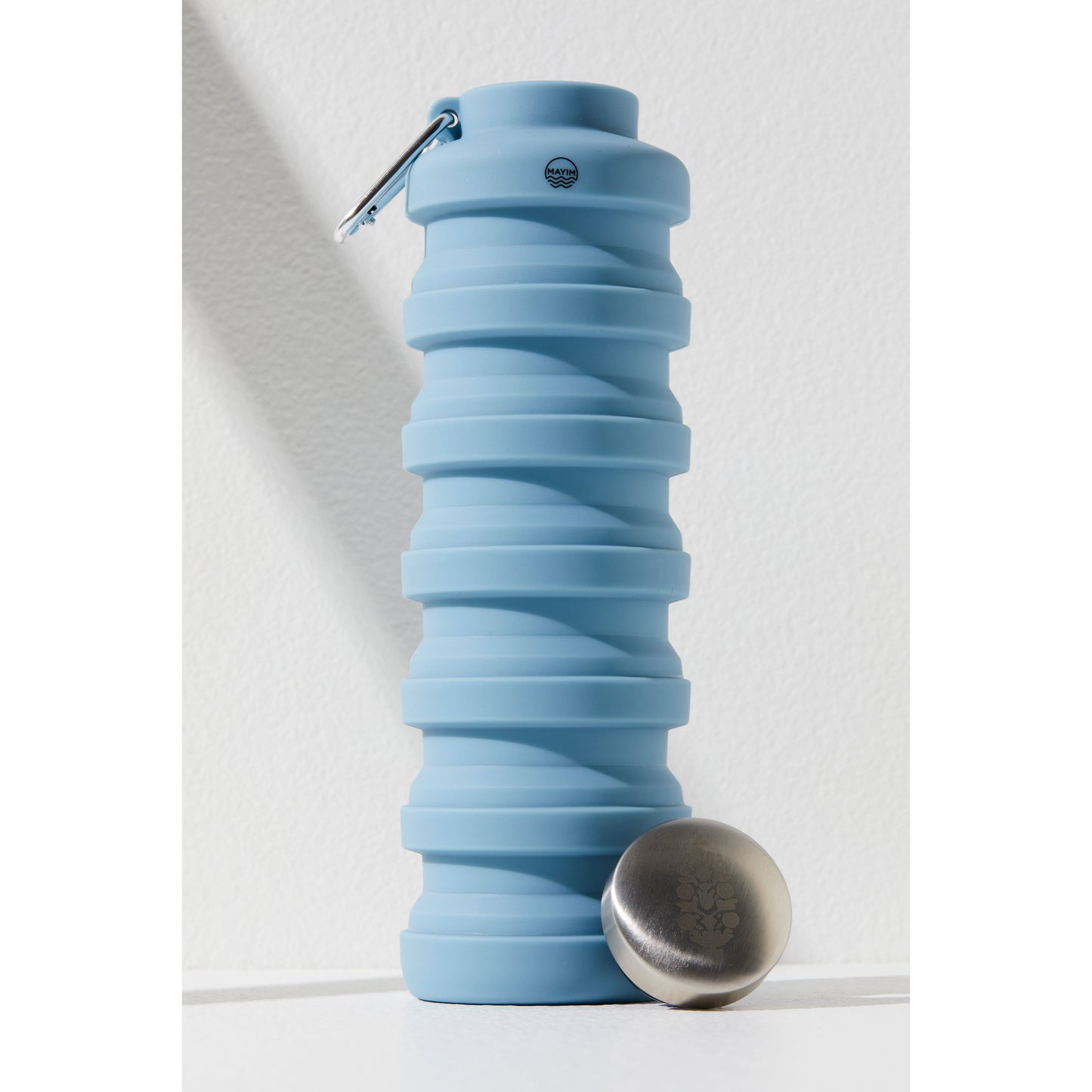 A collapsible, light blue silicone Free People Movement 24OZ Carabiner Bottle with its stainless steel lid placed next to it and a carabiner attached to its neck, designed as a tool for sustainable living.