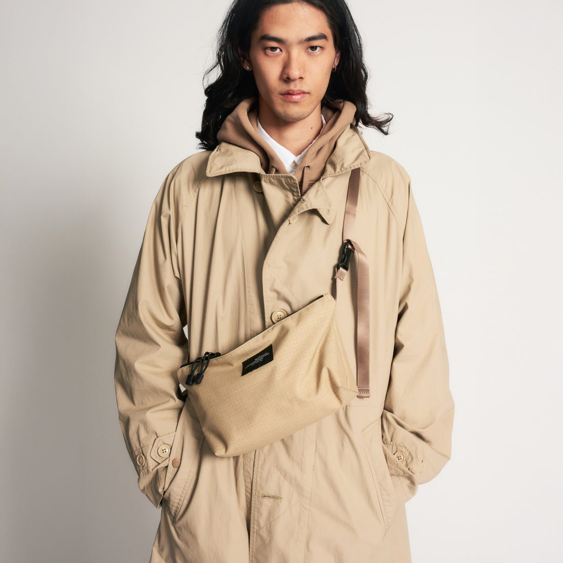 A person in a beige trench coat and hoodie stands against a plain background, wearing the Bags in Progress Fannypack Crossbody—a water repellent 3-way bag made from recycled polyester—across the chest.