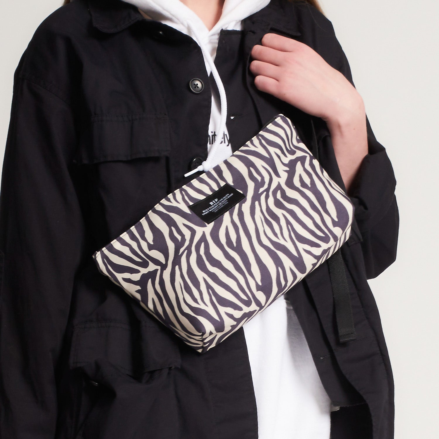 Person wearing a black jacket and white hoodie, carrying the Bags in Progress Fannypack Crossbody—a 3-way zebra print bag with a black label, made from recycled polyester—across their chest.