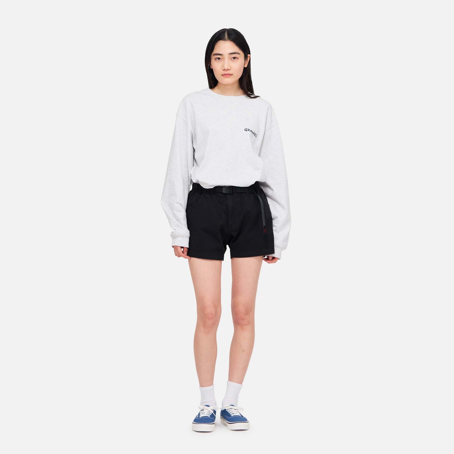 A woman stands facing forward in a light gray long-sleeve shirt and black Gramicci Very Short shorts, made from cotton stretch twill, paired with white socks and blue sneakers against a plain white background.