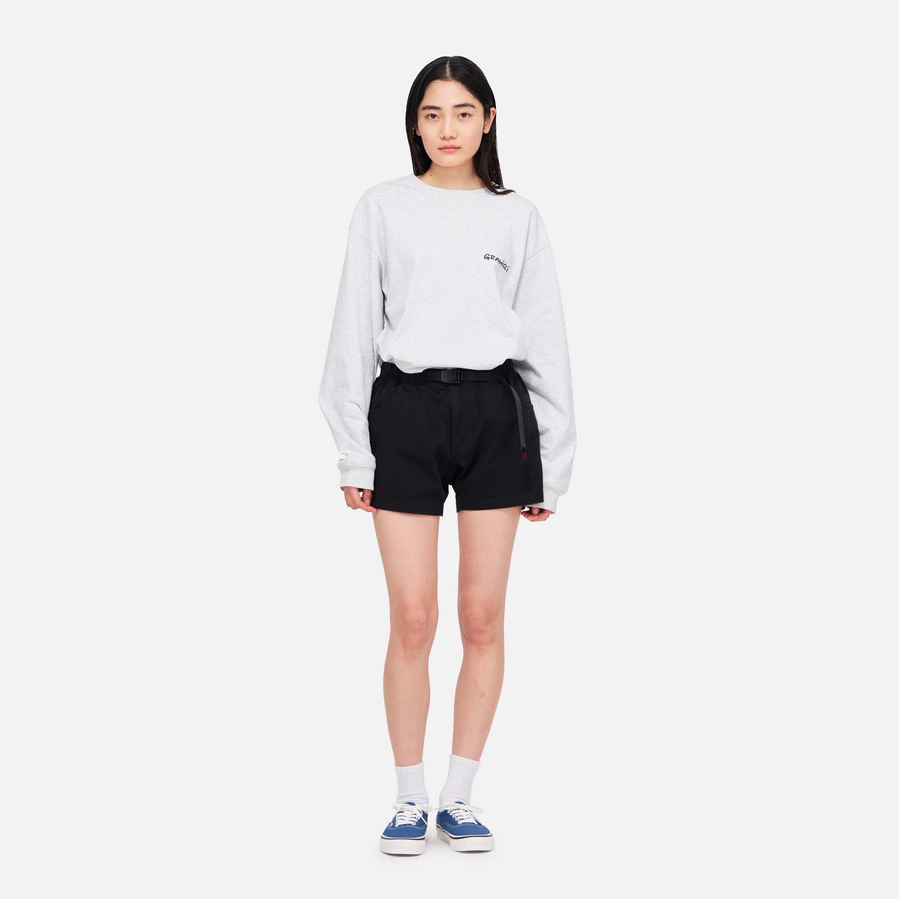 A woman stands facing forward in a light gray long-sleeve shirt and black Gramicci Very Short shorts, made from cotton stretch twill, paired with white socks and blue sneakers against a plain white background.