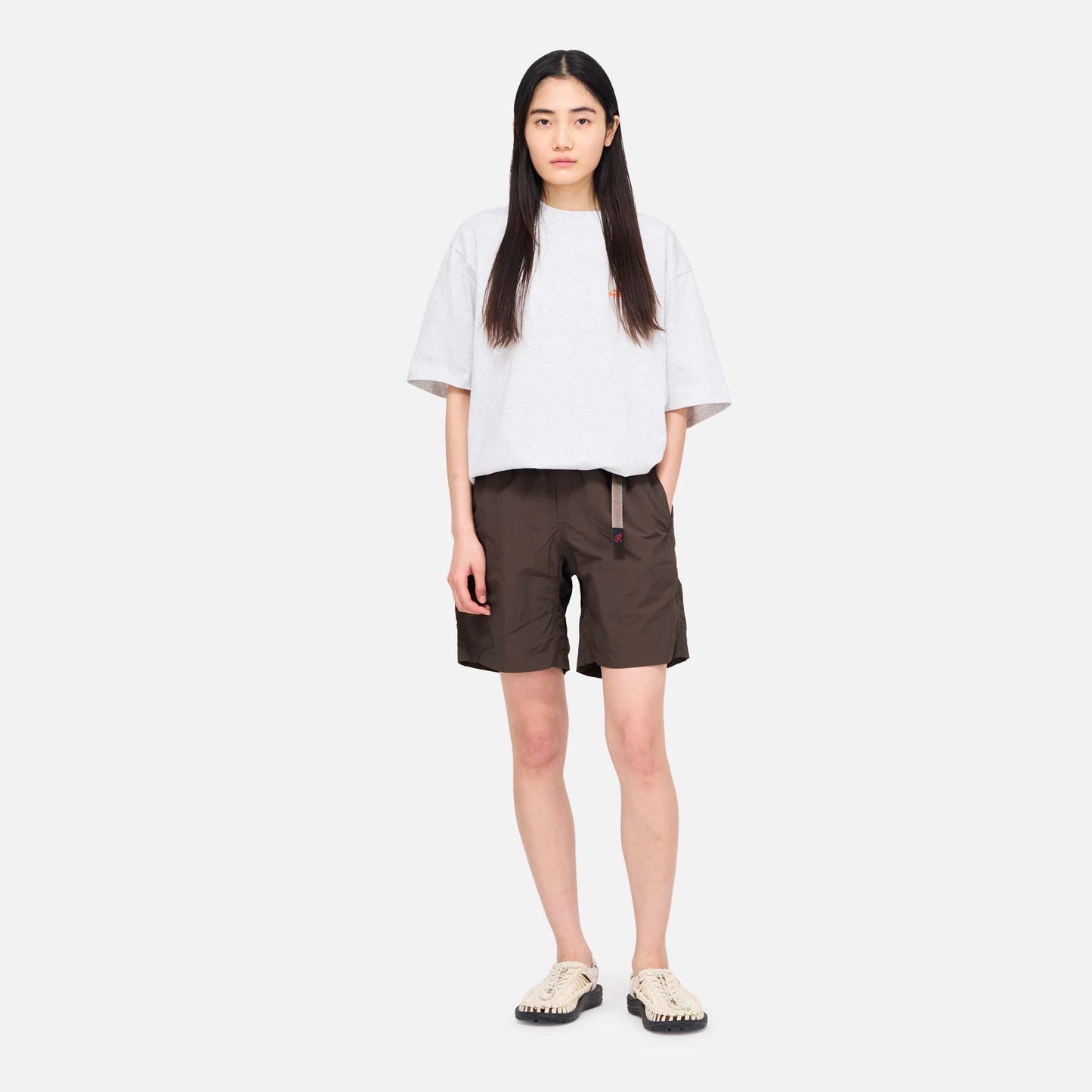 A woman stands facing forward in a loose white T-shirt, Gramicci Nylon Loose Short in dark brown, and beige sandals against a plain light background.