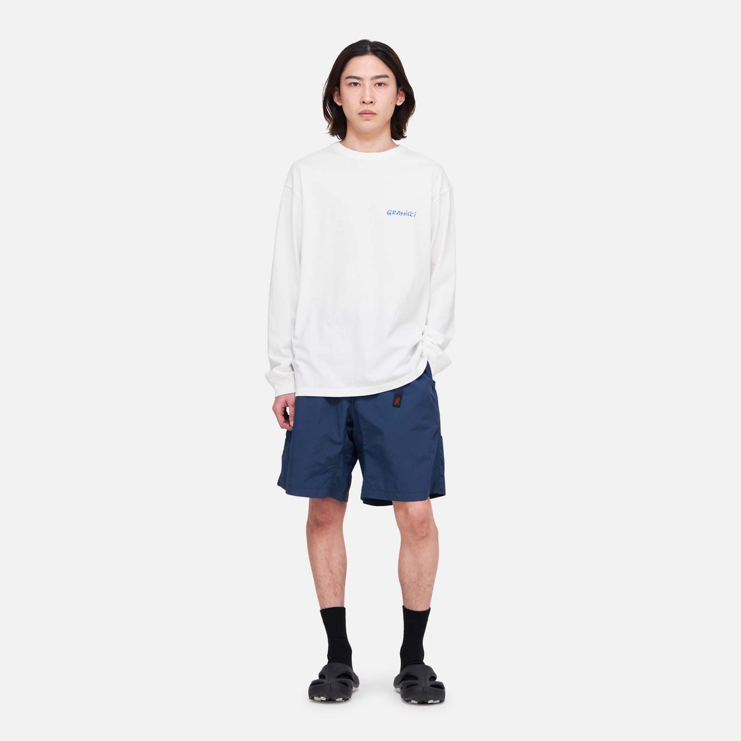 A person stands facing forward, wearing a white long-sleeve shirt and Gramicci's Nylon Loose Short in navy blue, which are water-repellent and wind-resistant, paired with black socks and sandals against a plain white background.