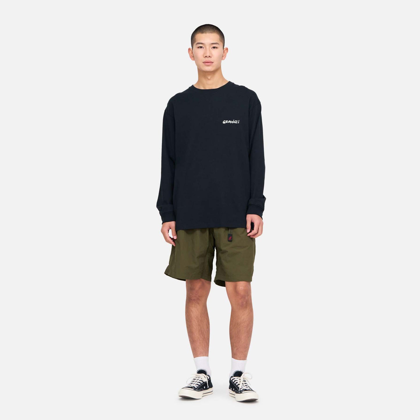 A person stands facing forward against a plain white background, wearing a black long-sleeve shirt, Gramicci Nylon Loose Shorts in olive green, white socks, and black sneakers.