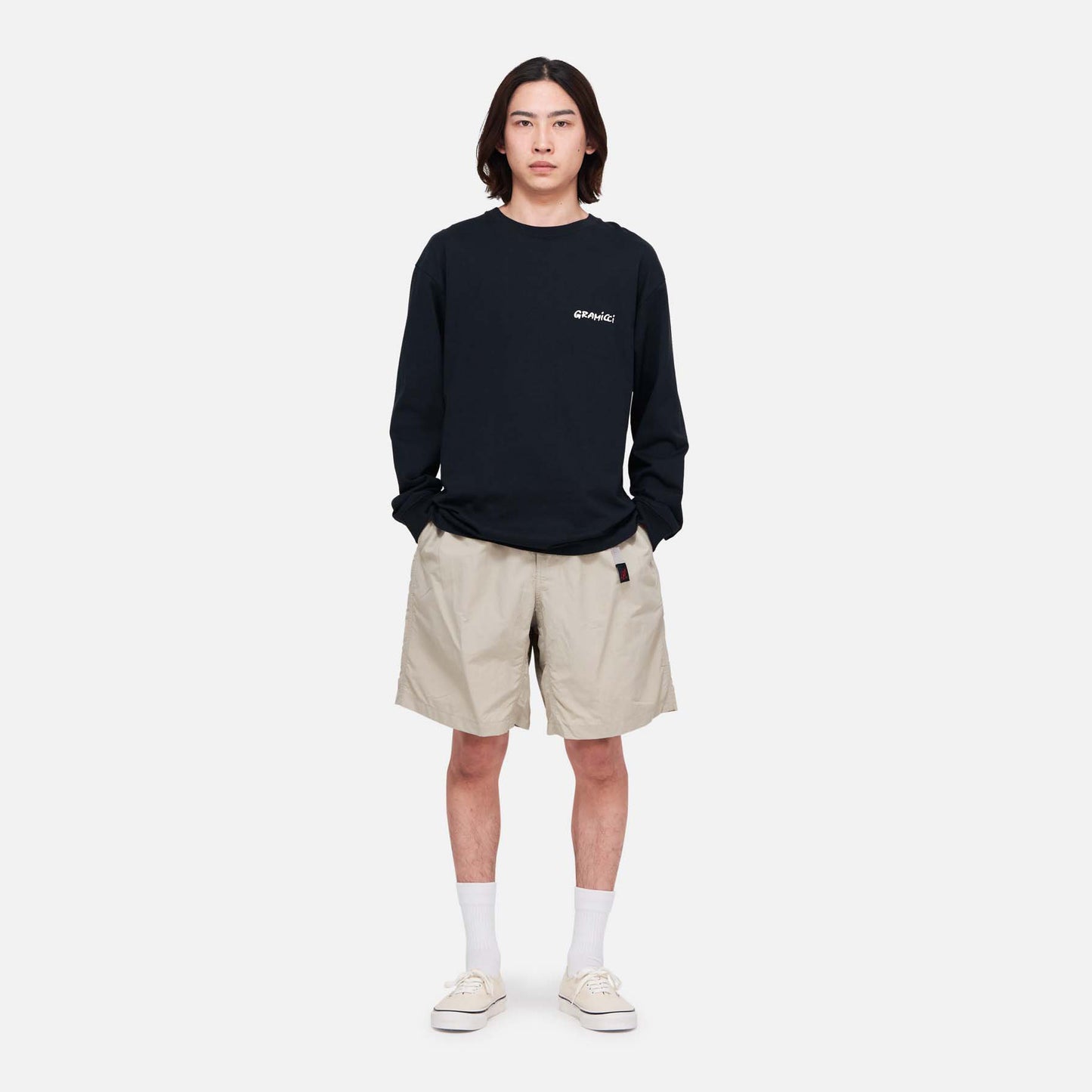 A person stands facing forward against a plain white background, wearing a black long-sleeve shirt, Gramicci Nylon Loose Short in wind-resistant beige nylon, white socks, and beige sneakers.