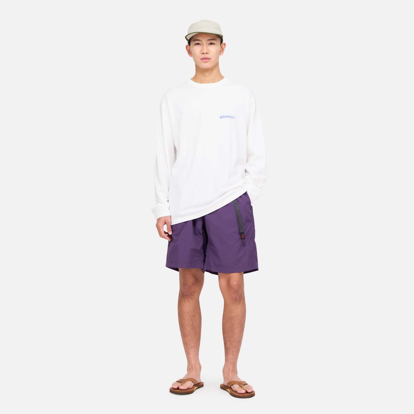 A person stands facing forward against a plain background, wearing a light-colored cap, white long-sleeve shirt, Gramicci Nylon Loose Short in purple, and brown sandals.