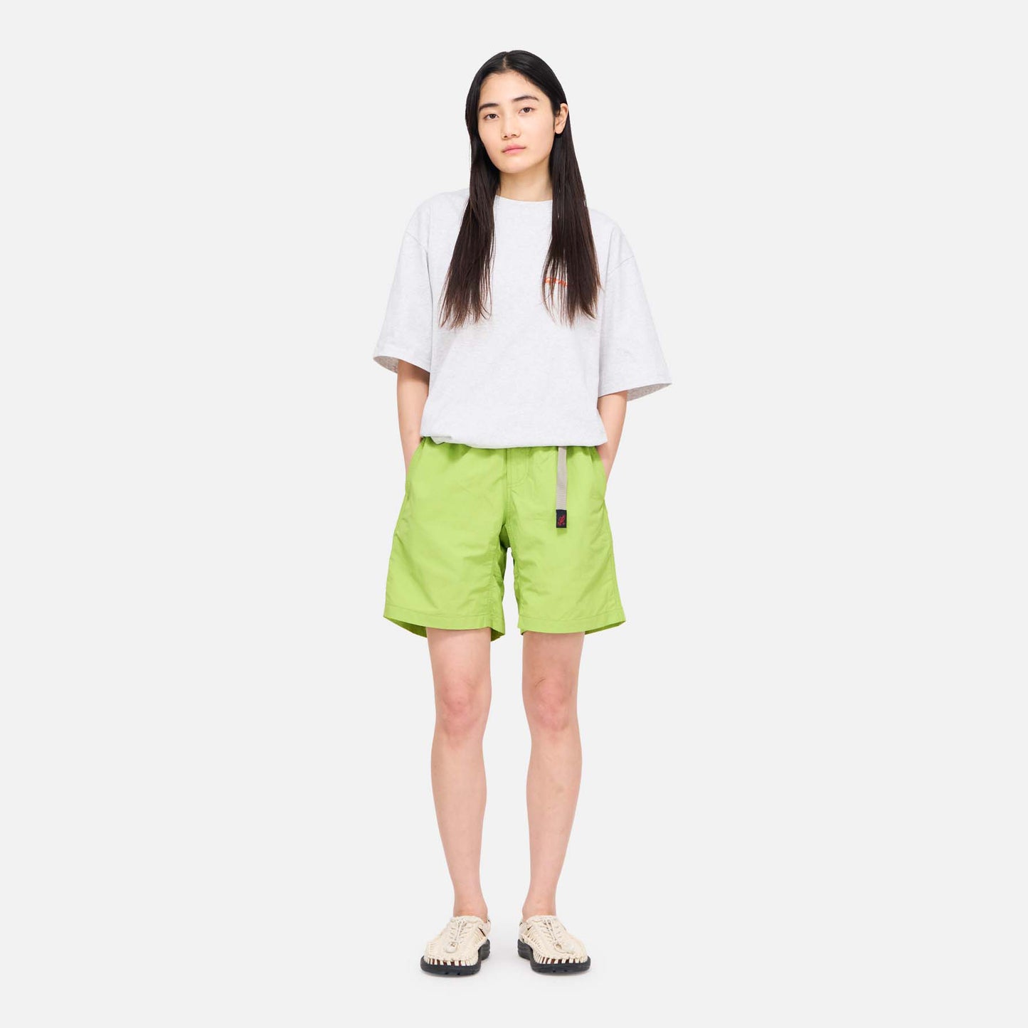 A person faces forward against a plain white background, wearing a loose white t-shirt, beige shoes, and Gramicci's Nylon Loose Short in bright green—water-repellent nylon shorts with a belt.