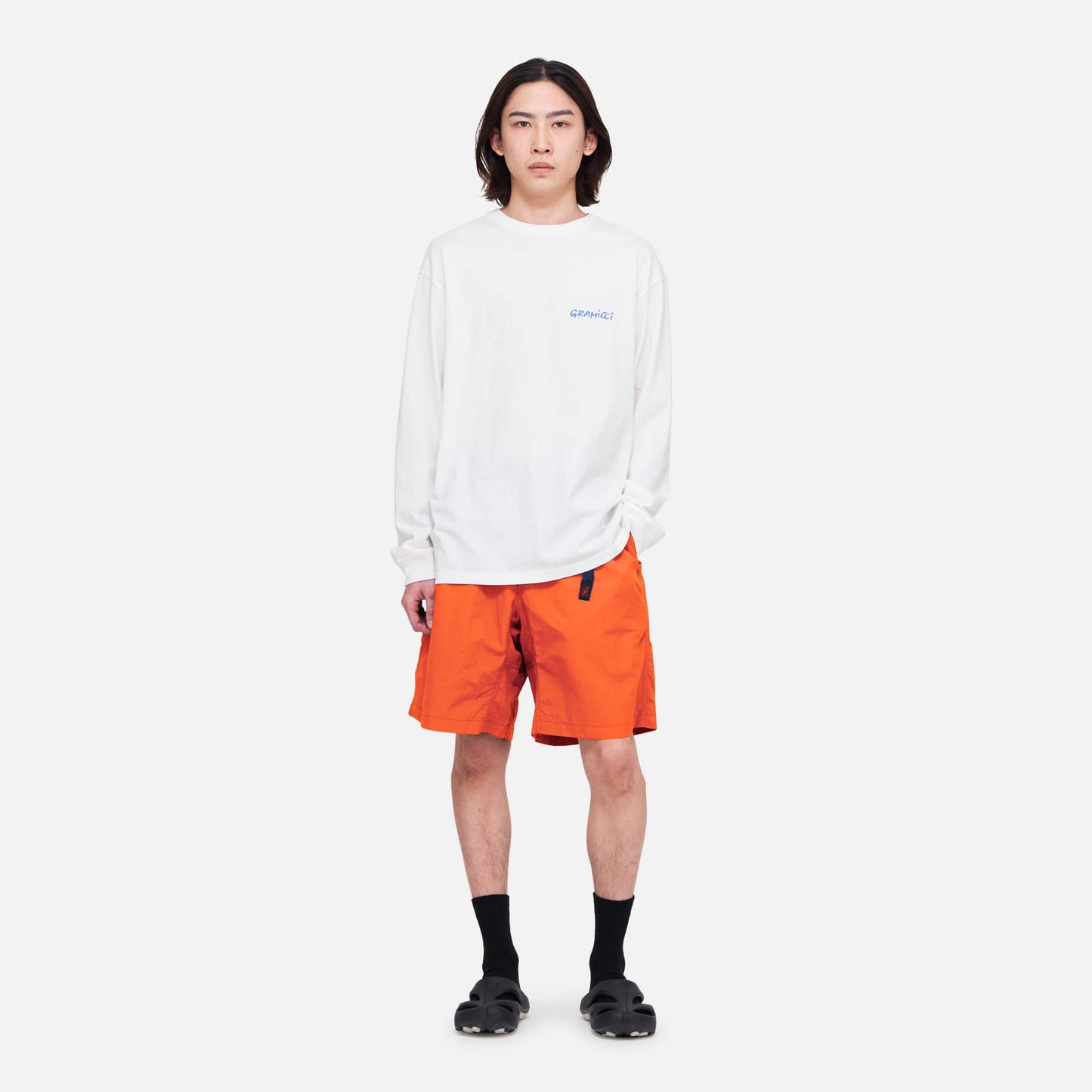 A person stands facing forward against a plain white background, wearing a white long-sleeve shirt, Gramicci Nylon Loose Short in bright orange, black socks, and black sandals.