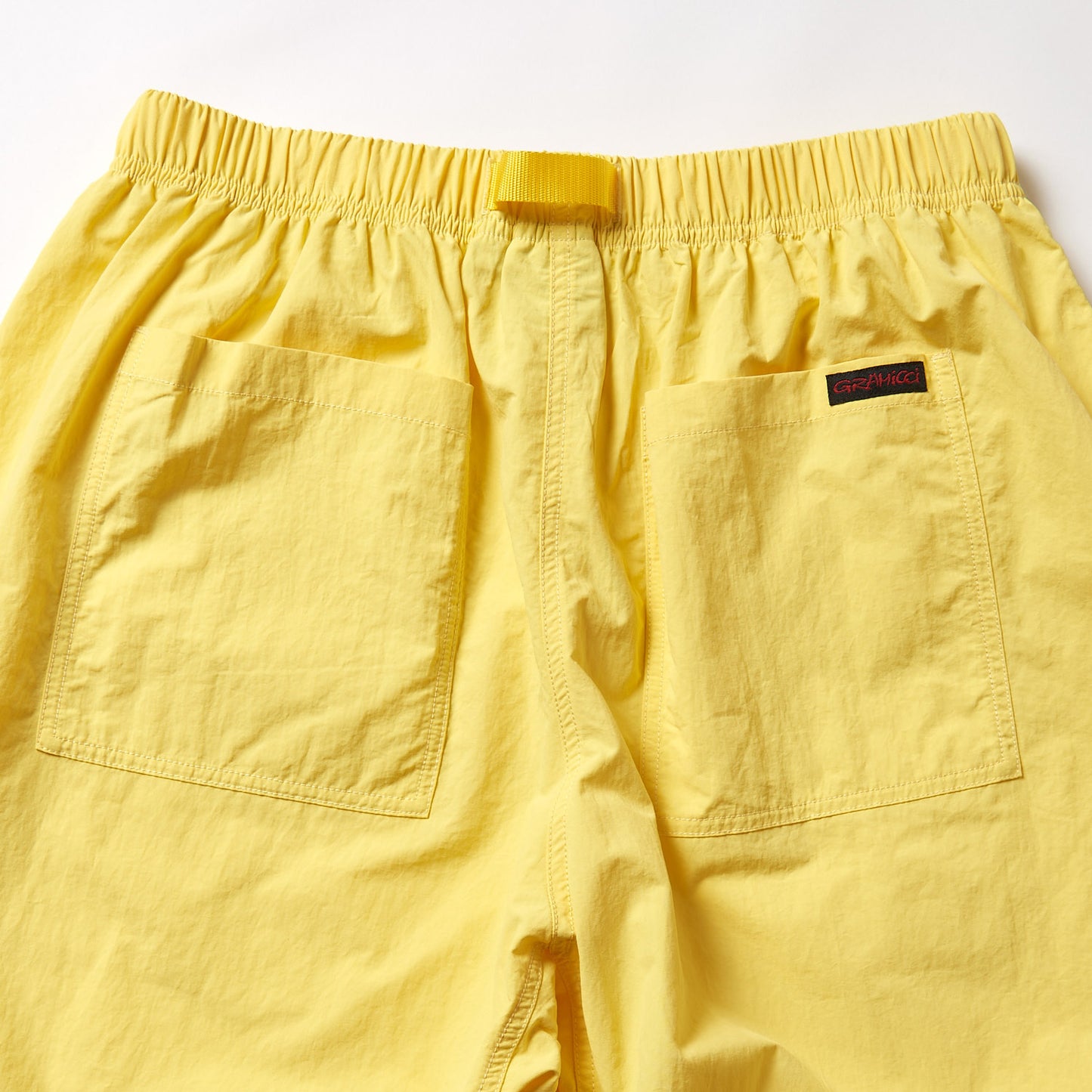 The Gramicci Nylon Loose Short features yellow nylon fabric, an elastic waistband, two large back pockets, a black and red label at the right waist, and is water-repellent for versatile wear.