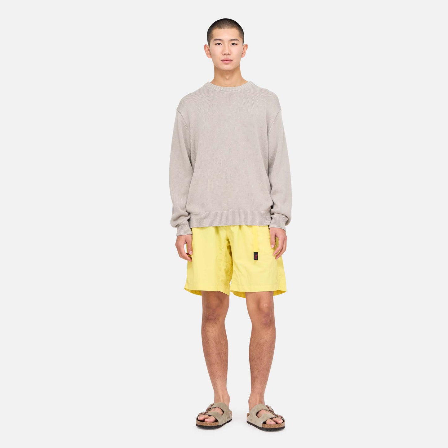 A person faces forward against a plain white background wearing a light grey sweater, Gramicci Nylon Loose Short in bright yellow (water-repellent and wind-resistant), and beige sandals.