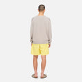 A person faces away, wearing a light gray sweater, Gramicci Nylon Loose Short in yellow water-repellent nylon, and sandals, posed against a plain white background.