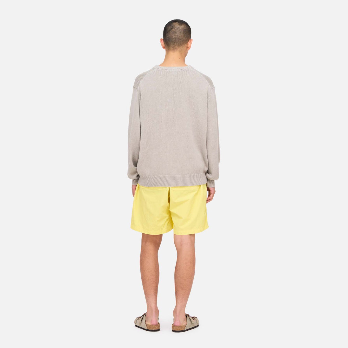 A person faces away, wearing a light gray sweater, Gramicci Nylon Loose Short in yellow water-repellent nylon, and sandals, posed against a plain white background.